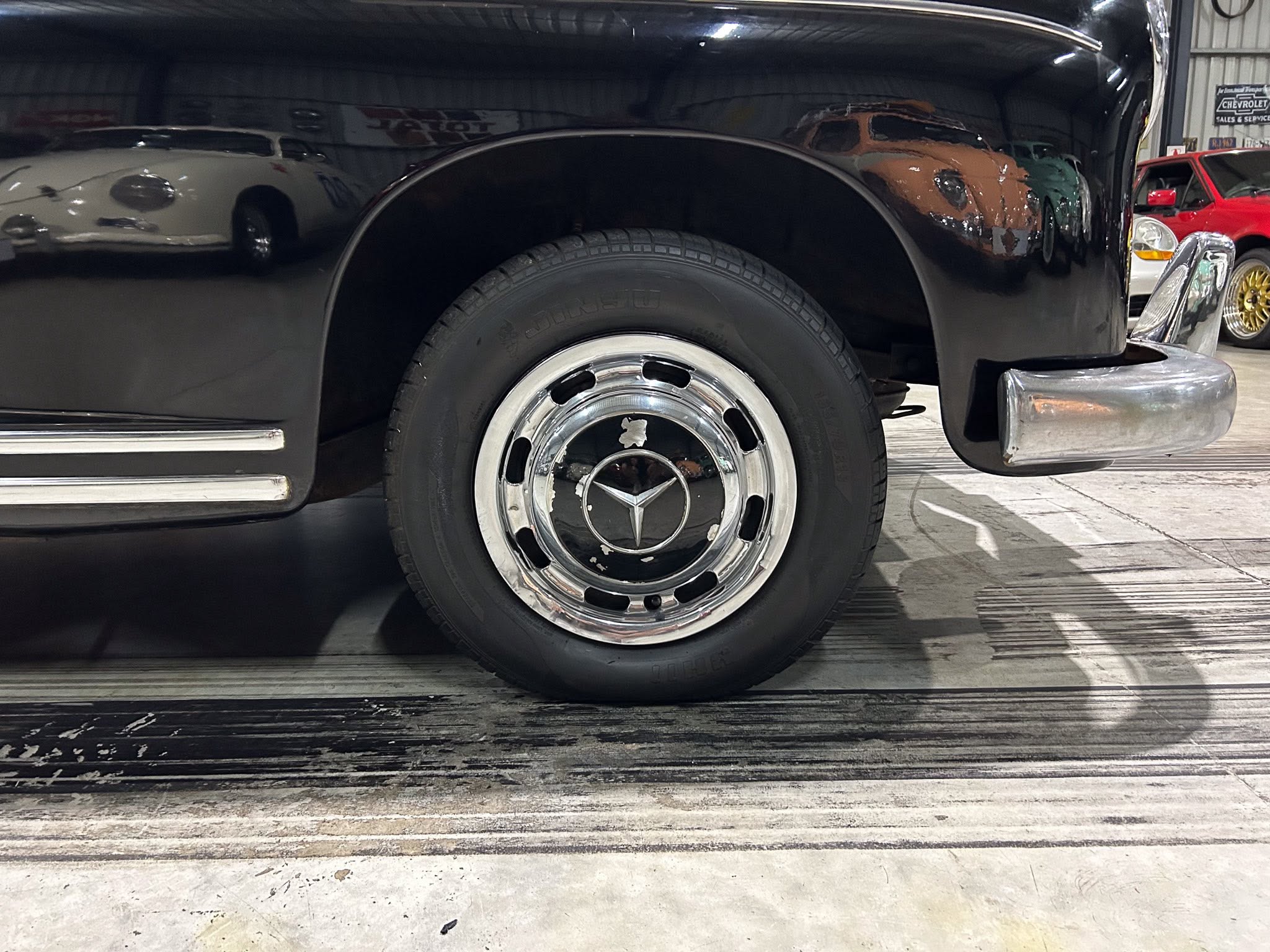 Close-up of a black vintage car’s front wheel and fender with chrome hubcap in a showroom reflection.