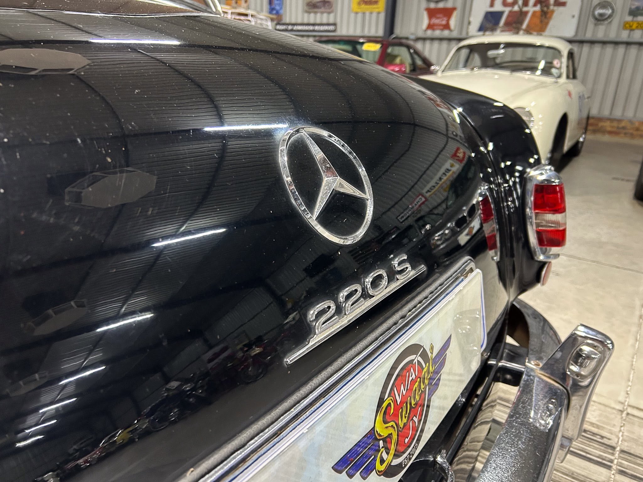 Close-up of the black Mercedes rear with chrome emblem, dust specks on the trunk, and vintage cars in a showroom.