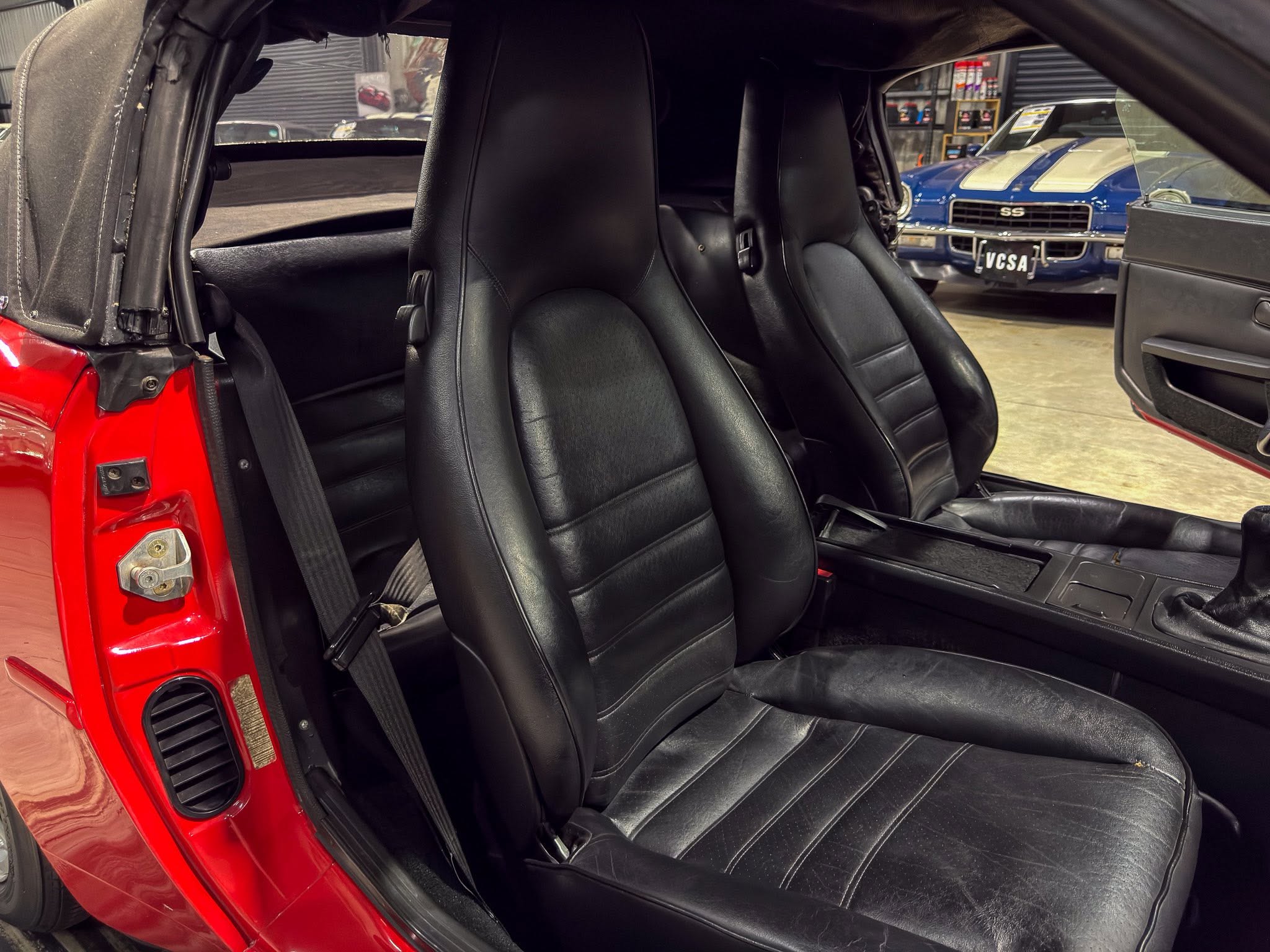 Red convertible interior with black leather bucket seats and an open door, shown in a showroom with a blue car in the background.