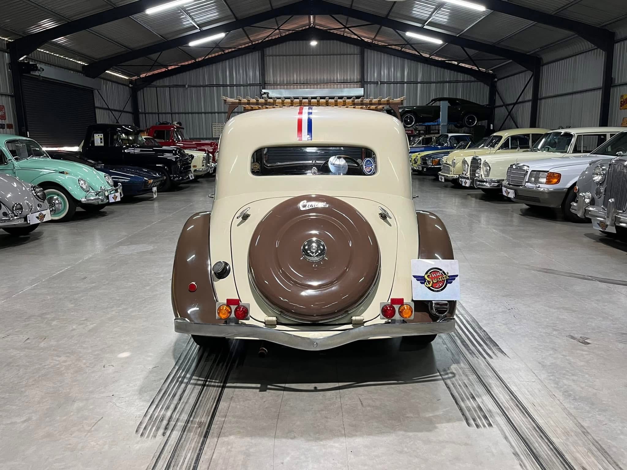 Rear view of a vintage beige car with a red, white, and blue stripe, inside a classic car showroom lined with numerous antique vehicles on both sides.