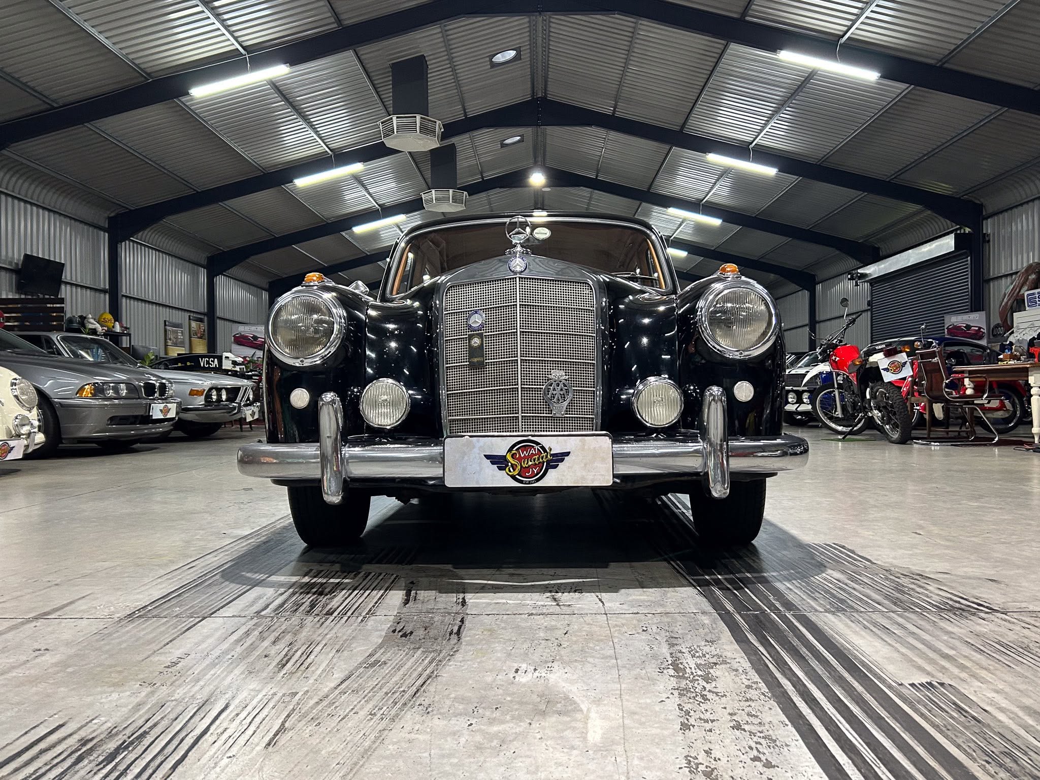 Front view of a vintage black luxury car inside a showroom, with other cars and motorcycles visible in the background.