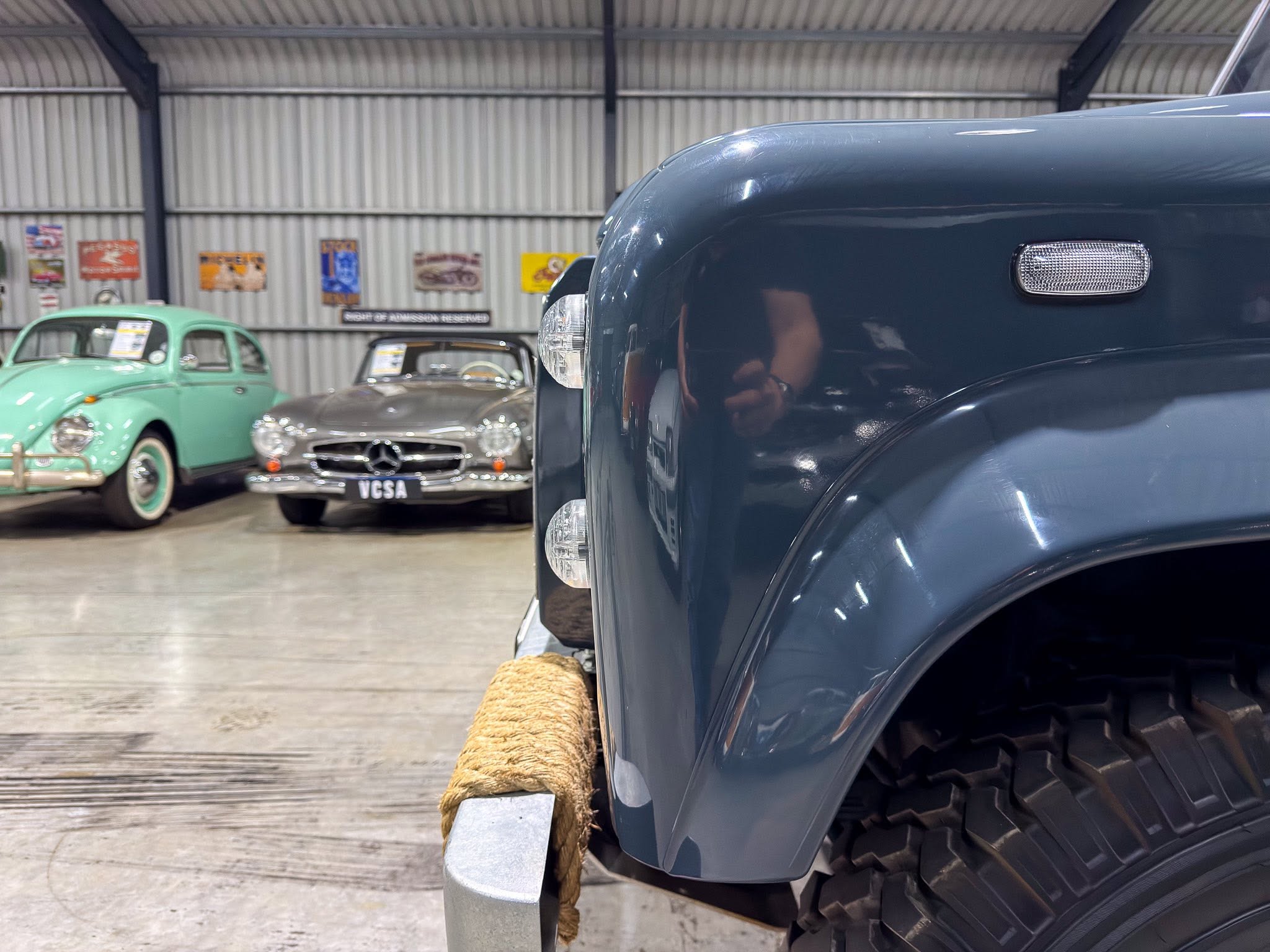 Close-up of a dark blue car's front fender with a rope tied to the bumper, inside a vintage car showroom with mint green and silver classics in the background.