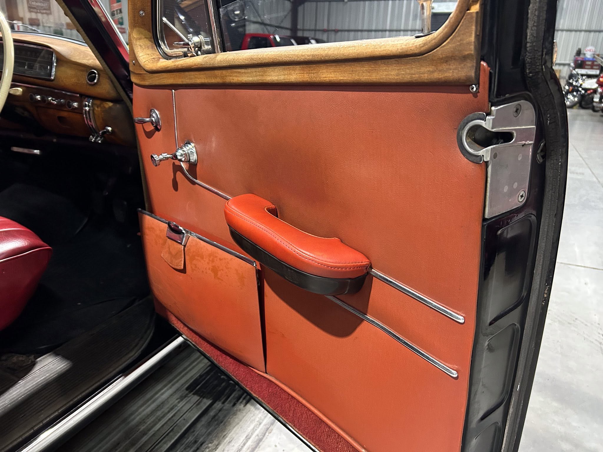 Close-up of a vintage car door interior featuring orange leather panel, chrome handles, and wood trim on top edge of the door