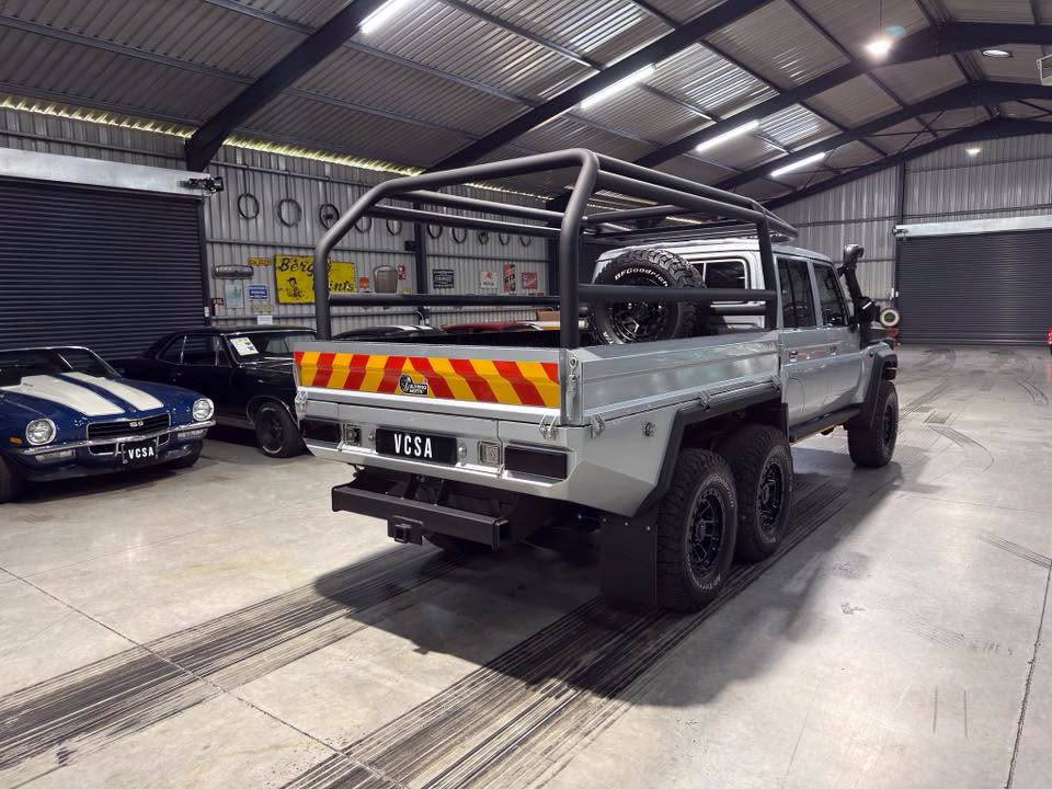 Grey dual-cab utility truck with a large tubular rack in a workshop, spare tire mounted behind cab, and red-yellow chevron stripes on tailgate in foreground.