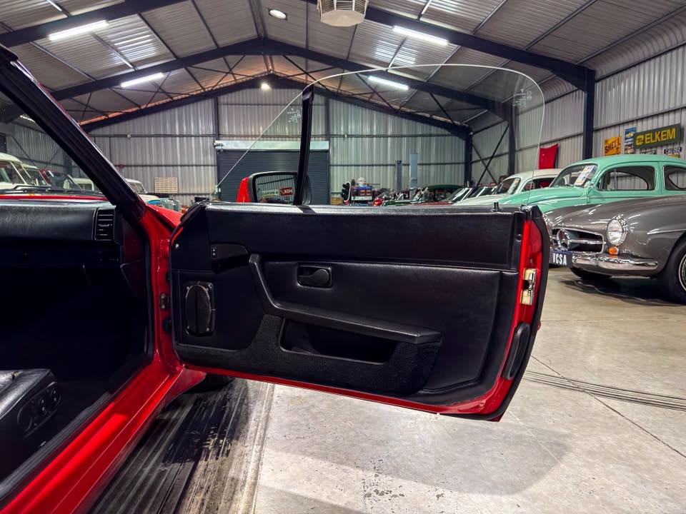 Red sports car with its driver-side door open, exposing a black interior, in a warehouse showroom with classic cars in the background.