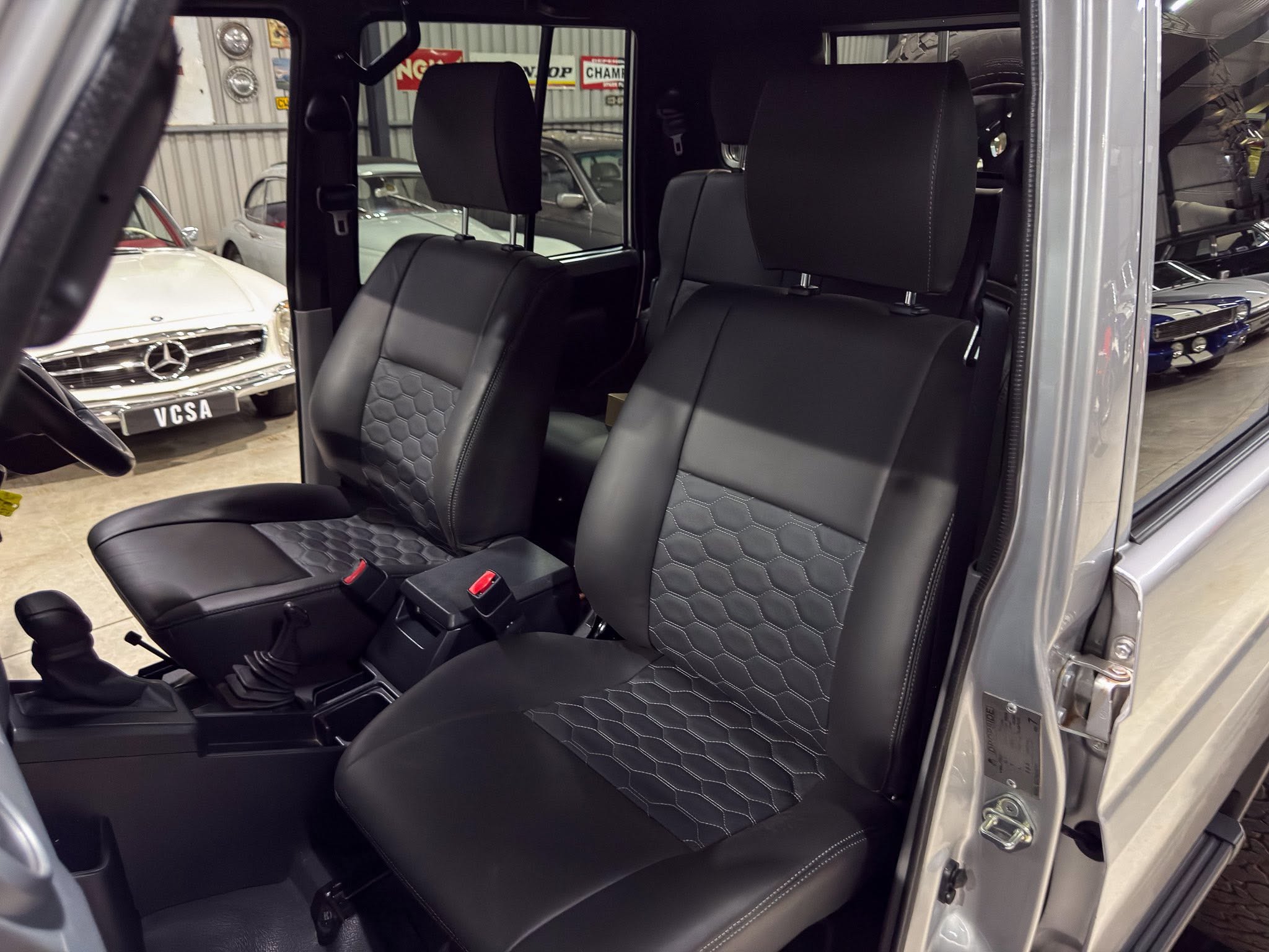 Interior of a gray SUV showing front seats in black quilted leather with hex stitching; driver's door is open in a showroom with vintage cars visible outside the window(s).