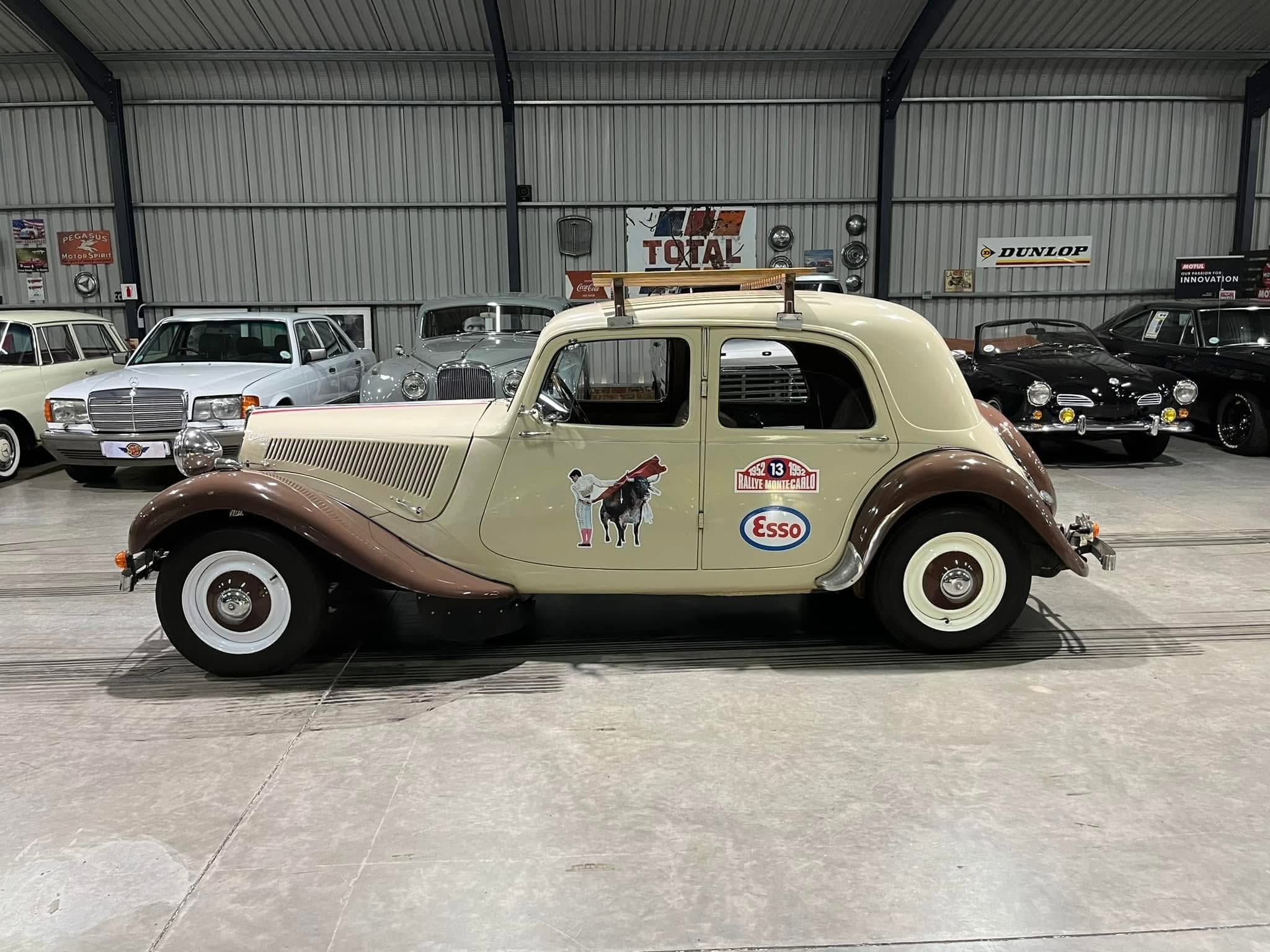 A row of vintage cars in a metal-walled showroom, with a beige classic car in the foreground featuring a wooden roof rack and retro decals.