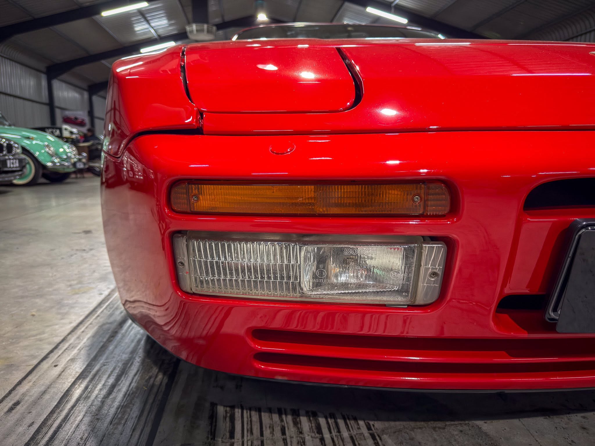 Close-up of a shiny red car front with an amber turn signal and rectangular headlight in an indoor showroom, other classic cars visible in the background