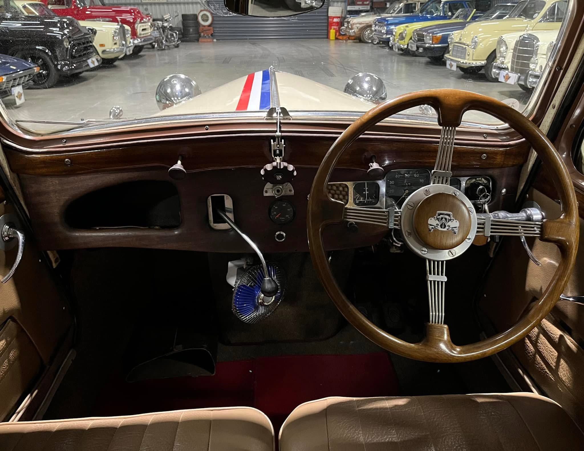 Driver's view of a vintage car interior with a wooden dashboard and steering wheel, showroom full of classic cars.
