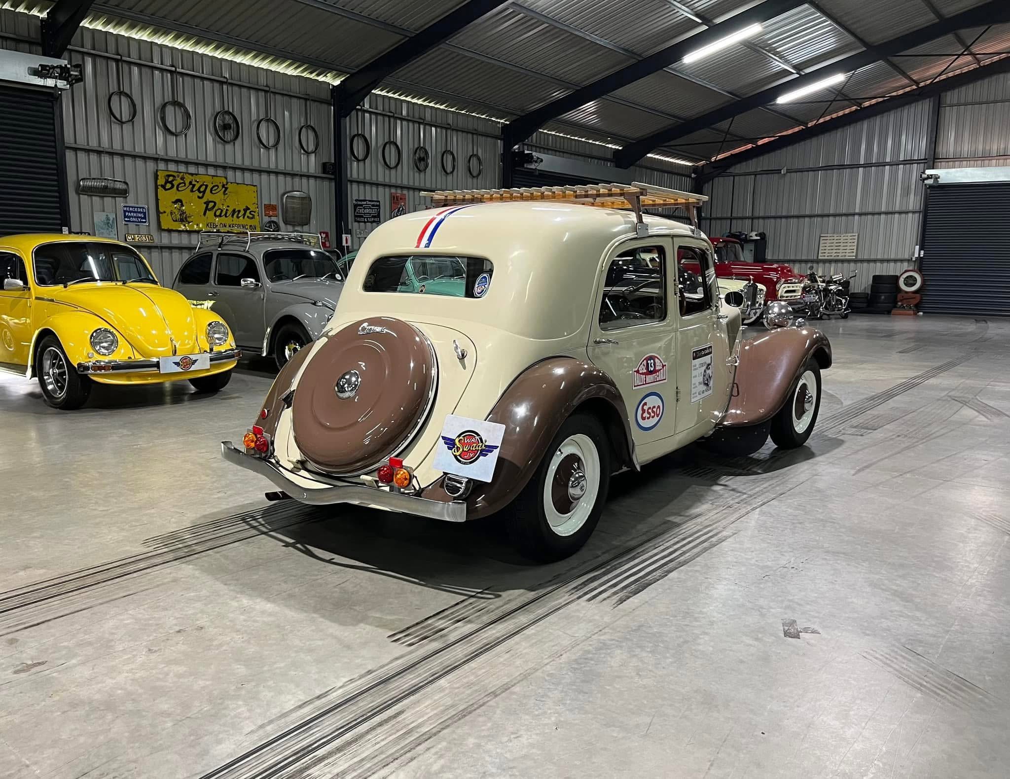 Indoor classic car showroom with a cream-and-brown vintage car in the foreground and a bright yellow car on the left, inside a metal-walled warehouse.