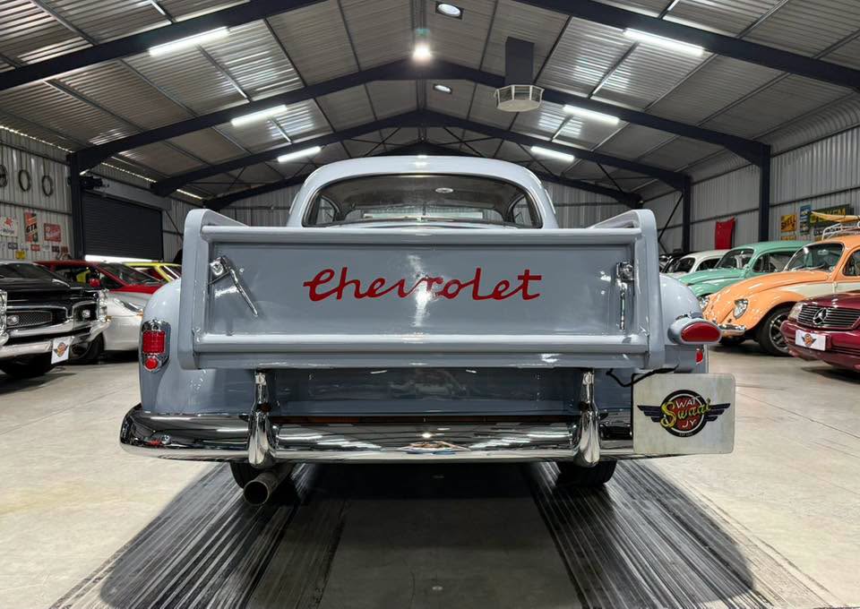 Rear view of a pale blue vintage Chevrolet pickup in a showroom, with red 'Chevrolet' script on the tailgate and a chrome bumper nearby with other classic cars in the background.
