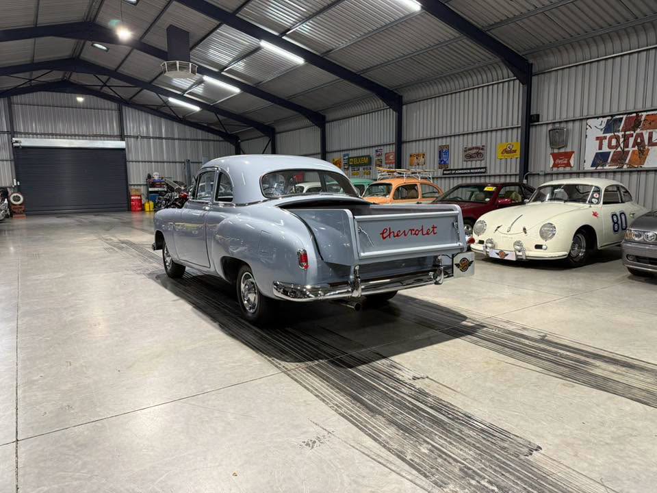 Classic light-blue vintage Chevrolet with open tailgate in a car showroom, other classic cars visible in background.