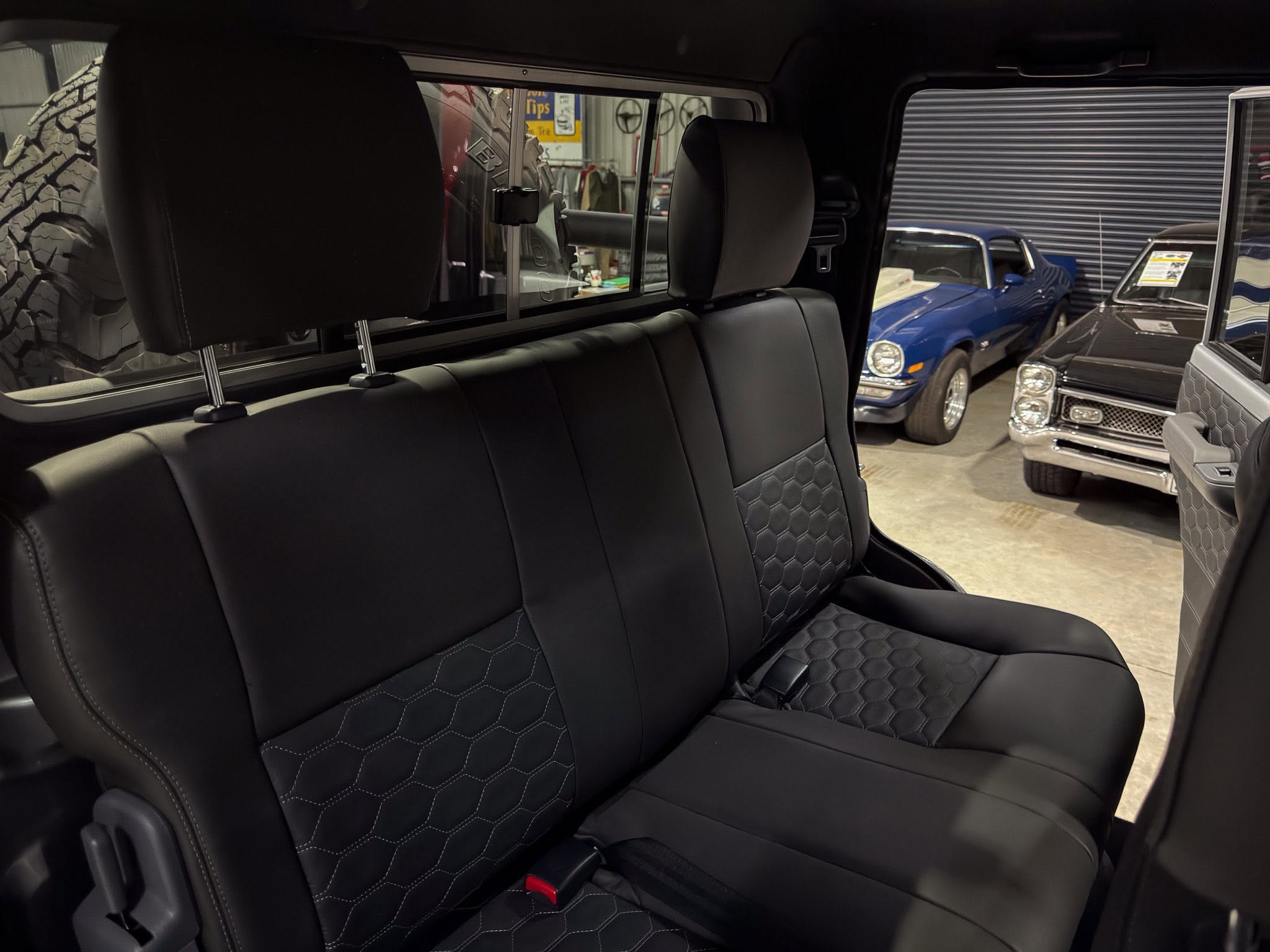 Backseat of a dark leather SUV with quilted stitching and headrests, inside a garage with classic cars visible outside the window.