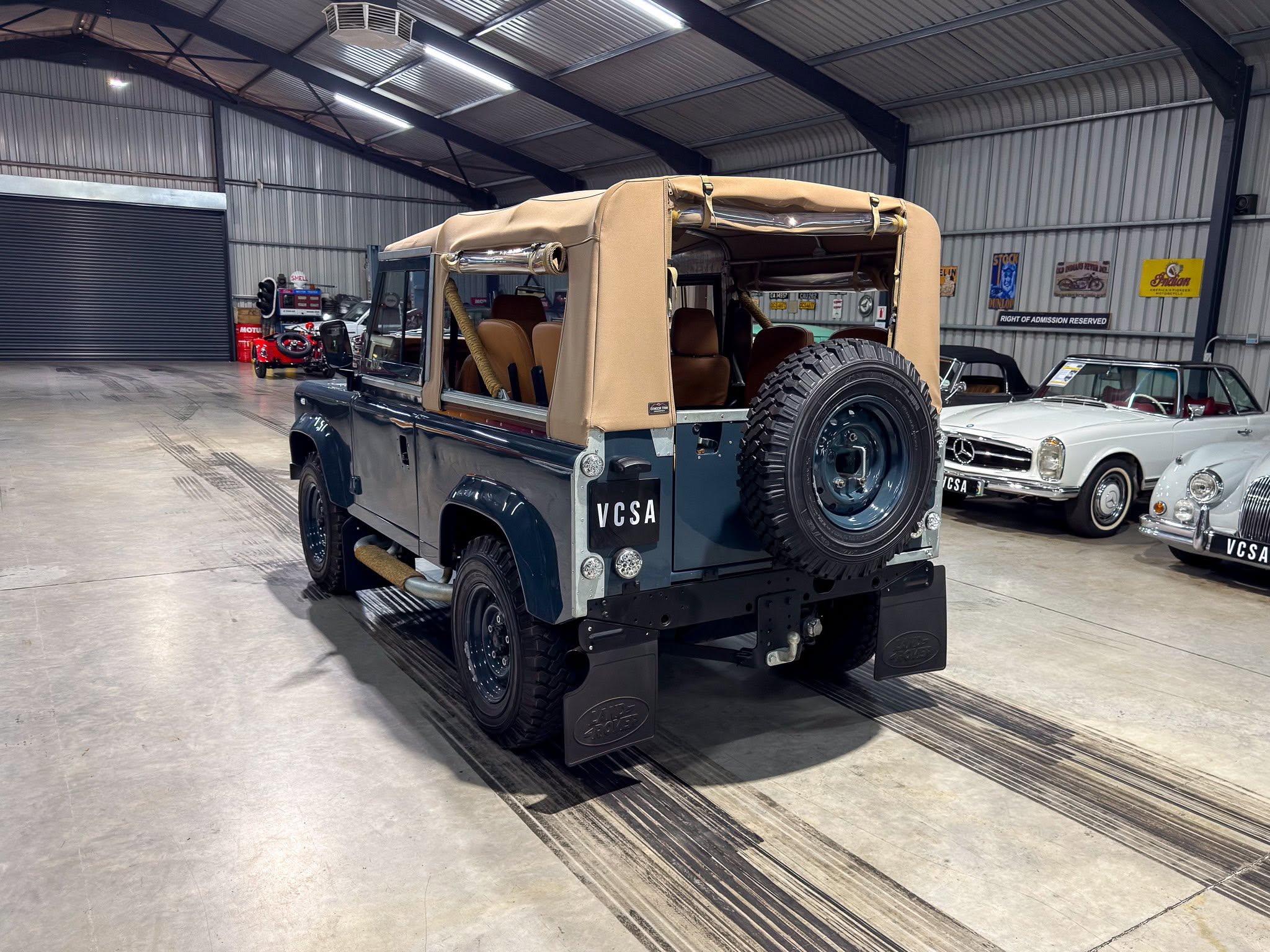 Inside a showroom-like garage with a dark blue vintage Land Rover Defender rear, tan soft top, spare tire mounted at back, and other classic cars visible in background.