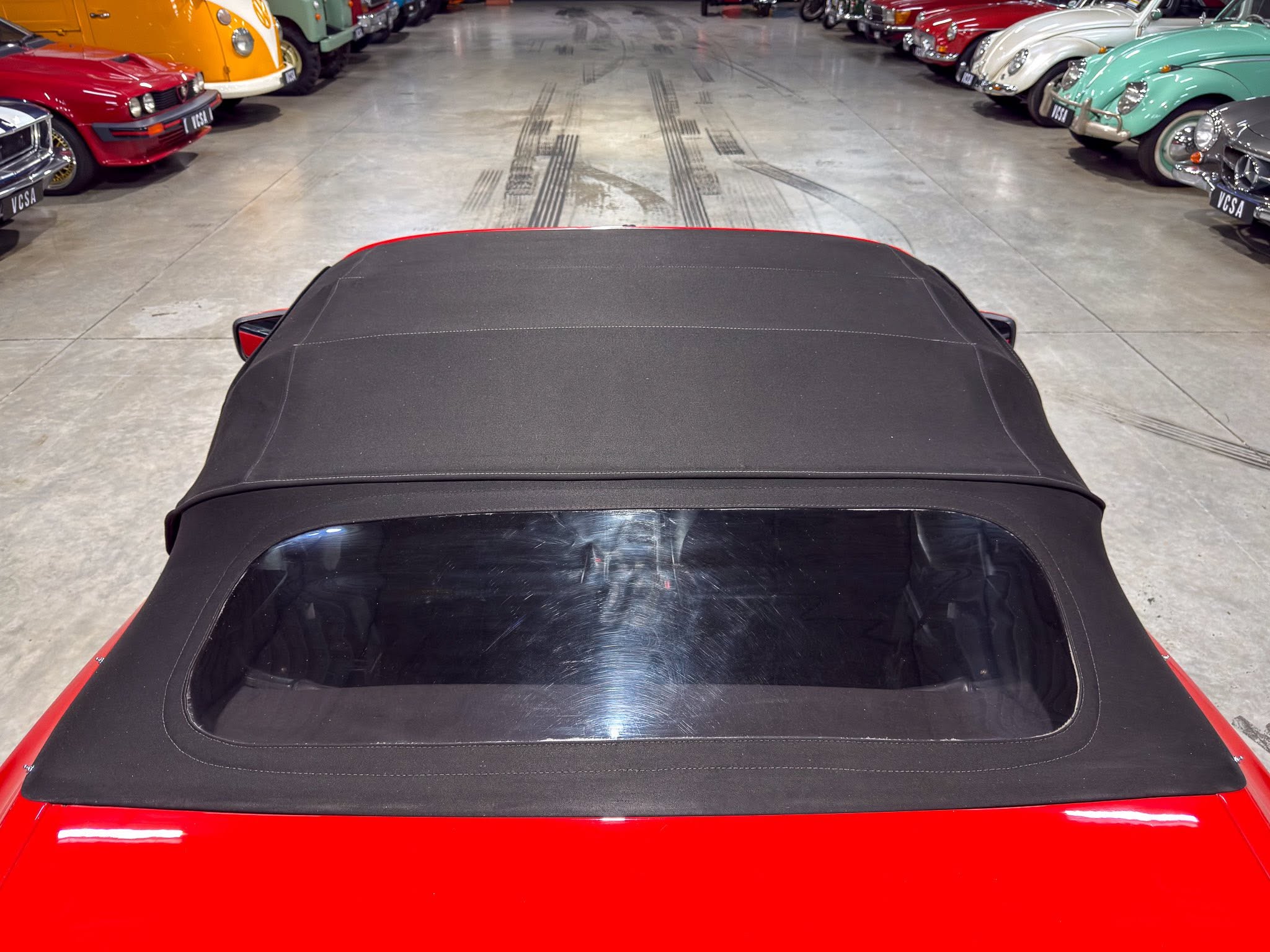 Photo taken from behind a red convertible with a black soft top, looking down a showroom-like garage lined with colorful vintage cars on both sides.