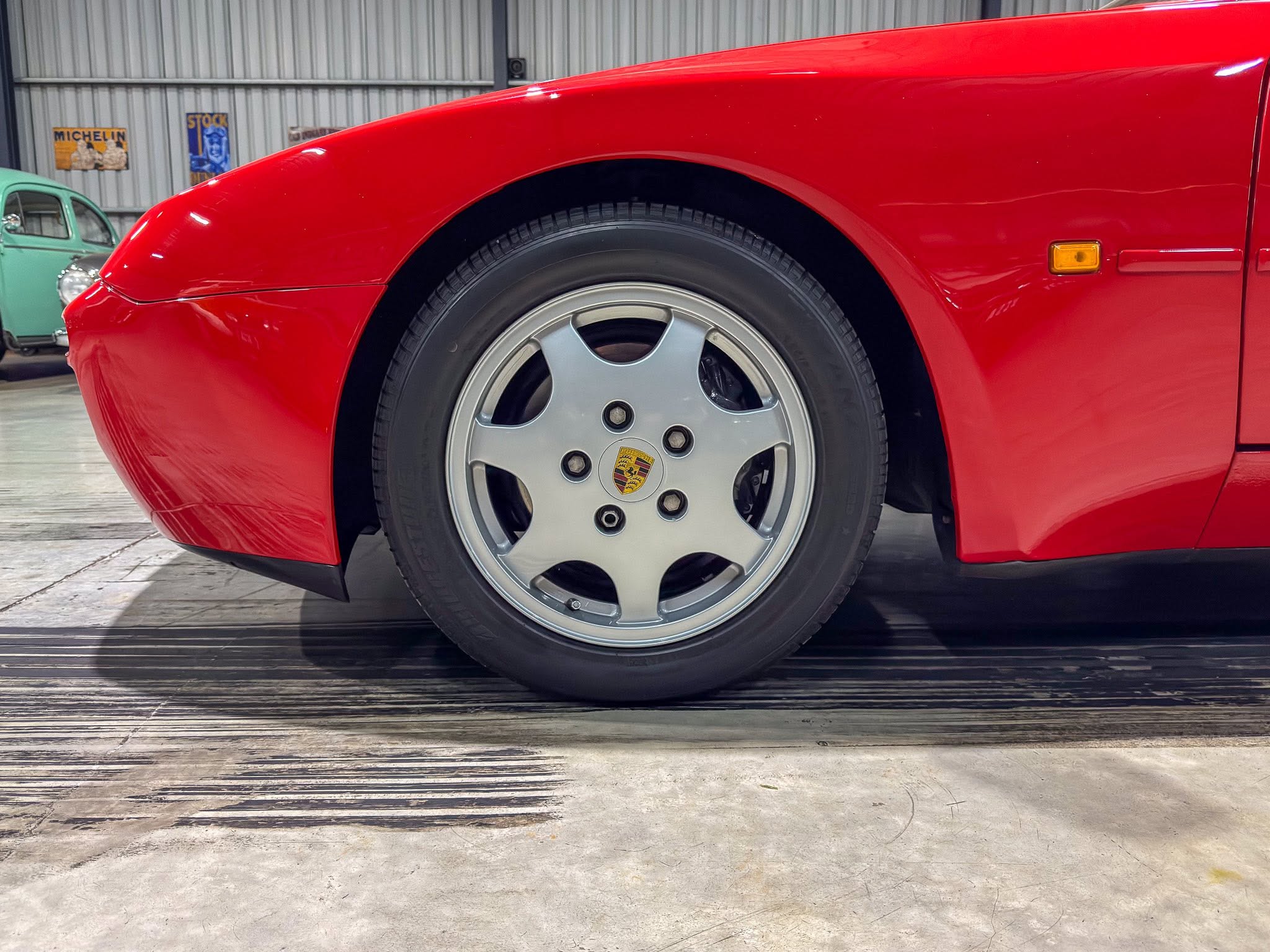 Front quarter and wheel of a red sports car in a garage, Porsche emblem on the alloy wheel with another vintage car in the background
