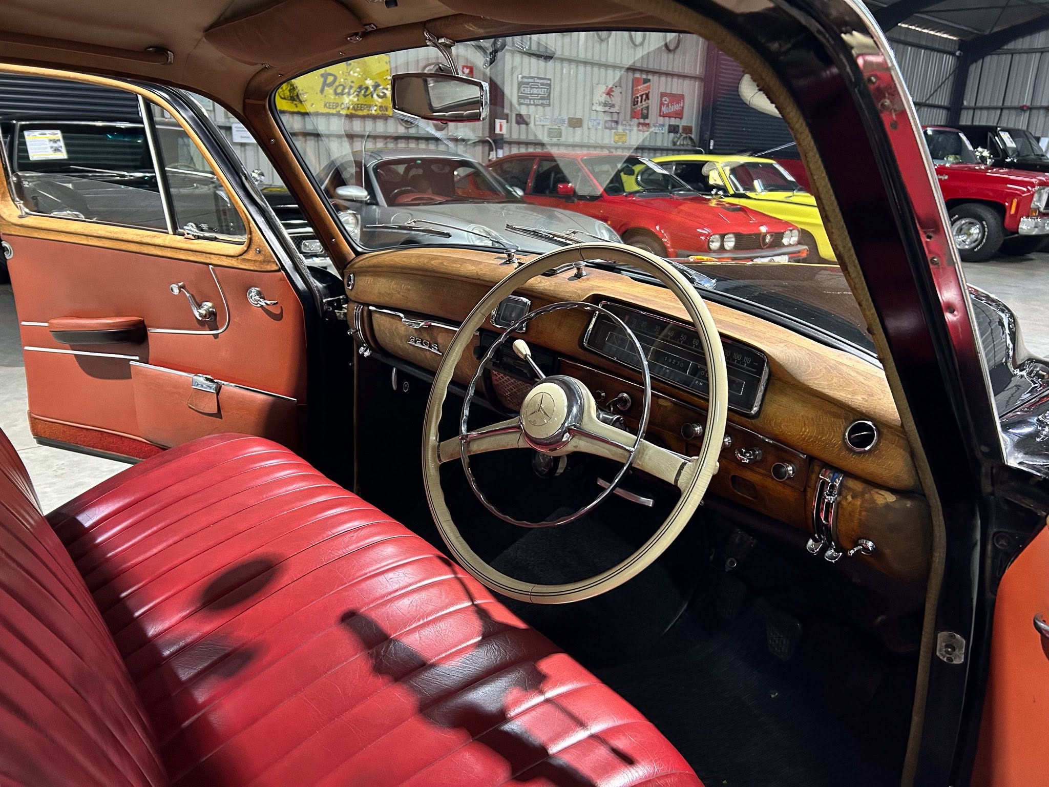 Interior of a vintage car with red leather seats, wood dashboard, and a classic steering wheel; other classic cars visible in a showroom background