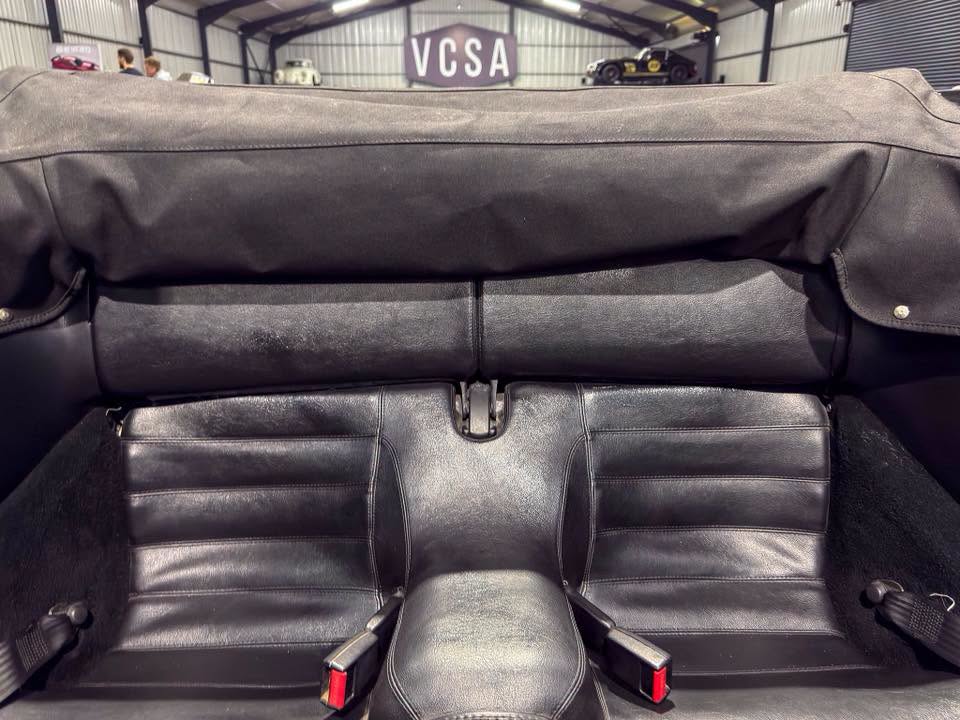 Back seats of a black convertible with leather upholstery, center console, and seat belts in a showroom with a VCSA sign in the background.
