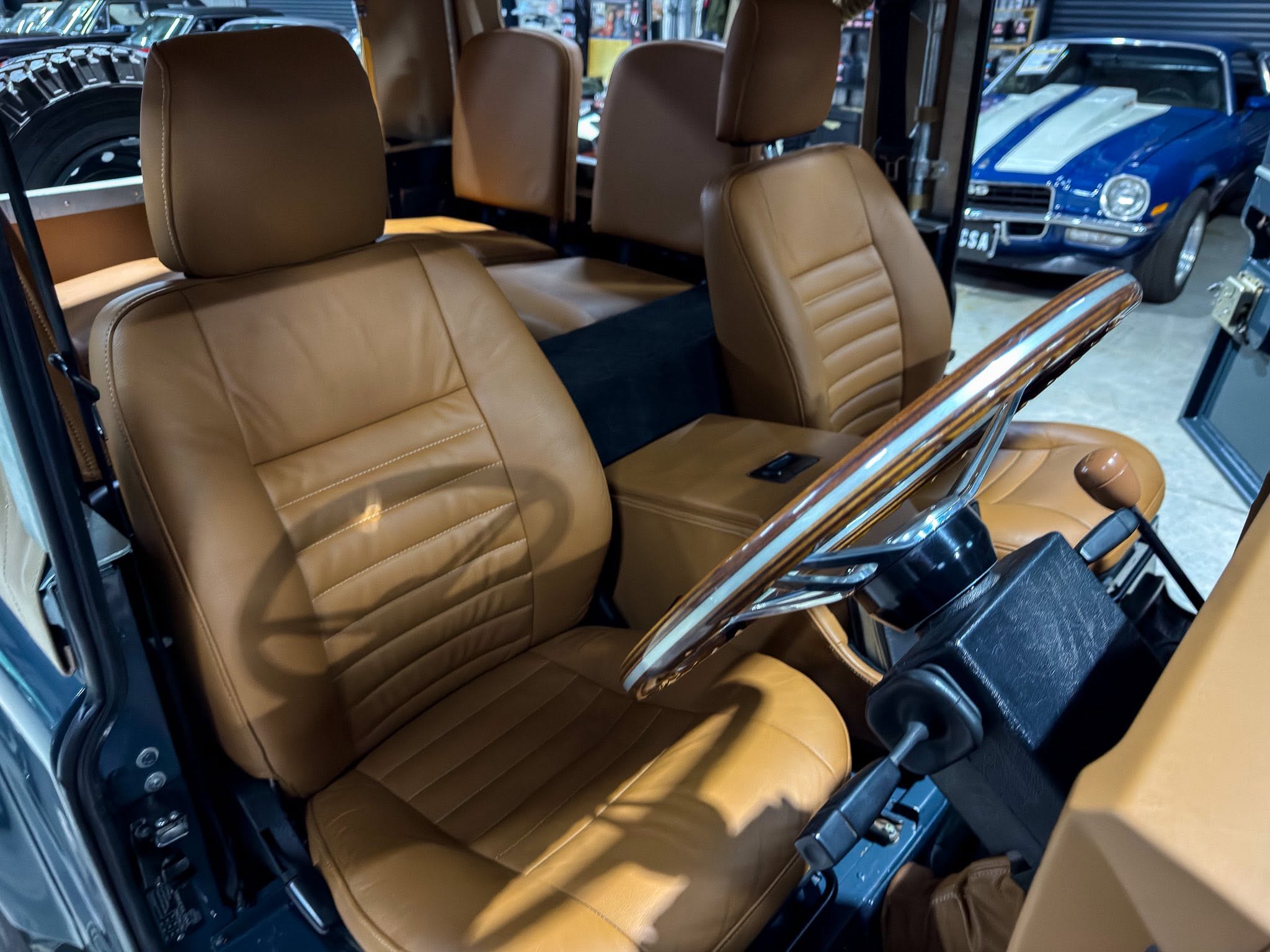 Tan leather car seats inside a vintage vehicle, with wood trim along the door and glossy black interior panels visible inside a showroom or garage.