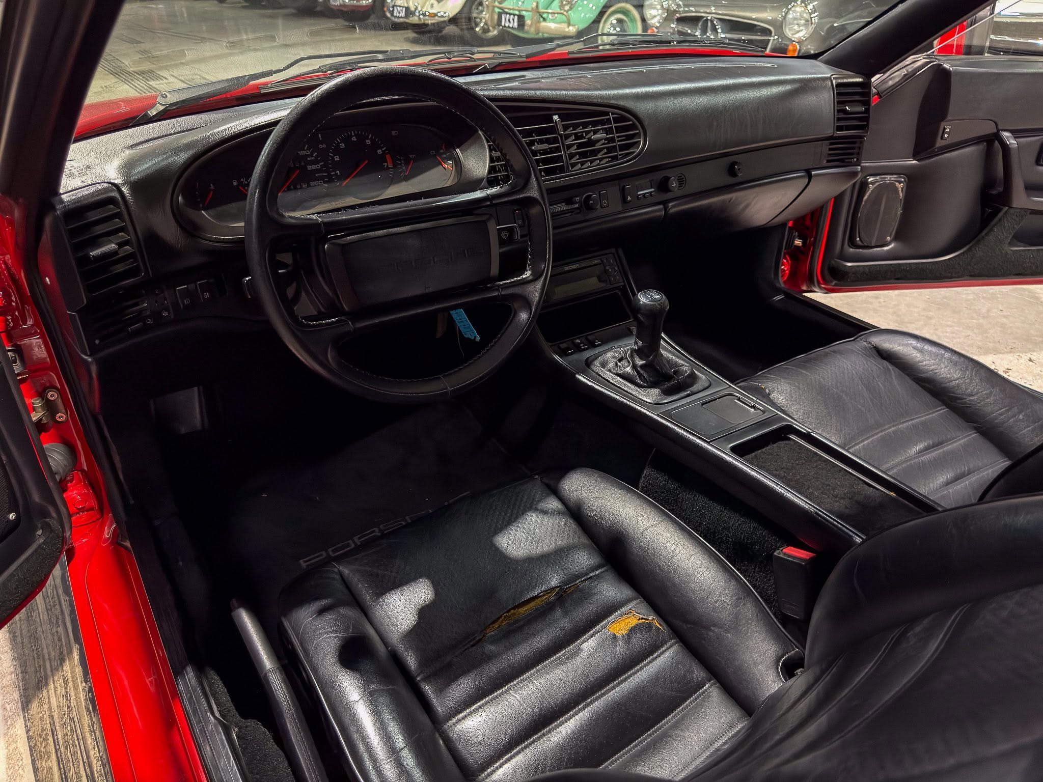 Red exterior of a Porsche seen through an open door, showing the black leather interior with steering wheel, gear shifter, and dashboard gauges.