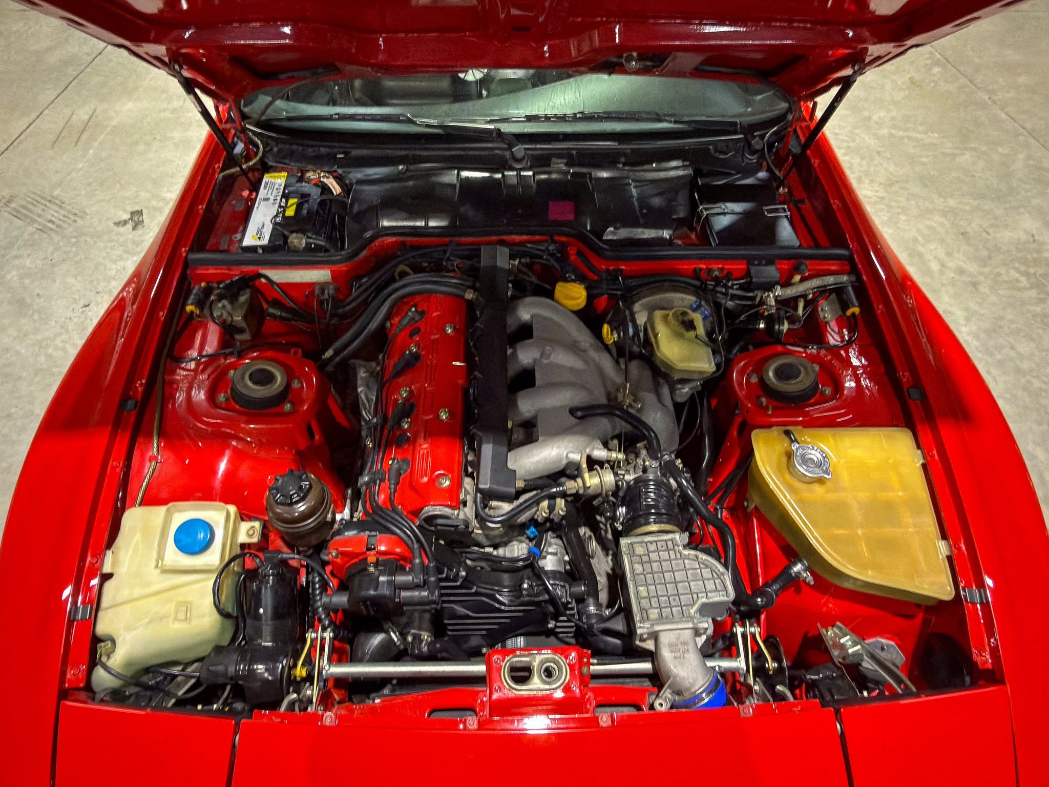 Red car with its hood open, showing the engine bay and various hoses, wires, and reservoirs visible from above.