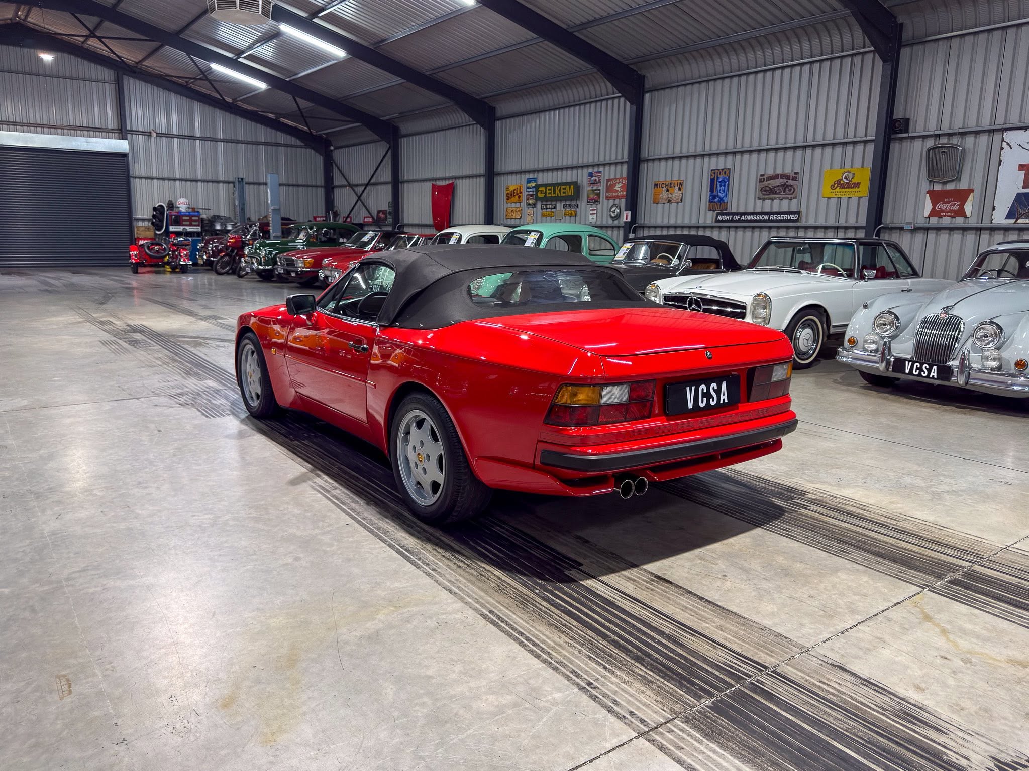 Red convertible sports car in a classic car showroom with vintage cars lined along a corrugated metal wall and retro signs above.