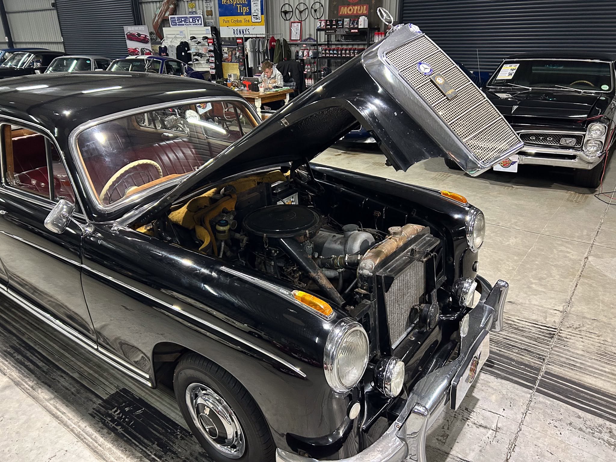 Close-up of a vintage black car with the hood open, engine visible in a workshop with other classic cars in the background.