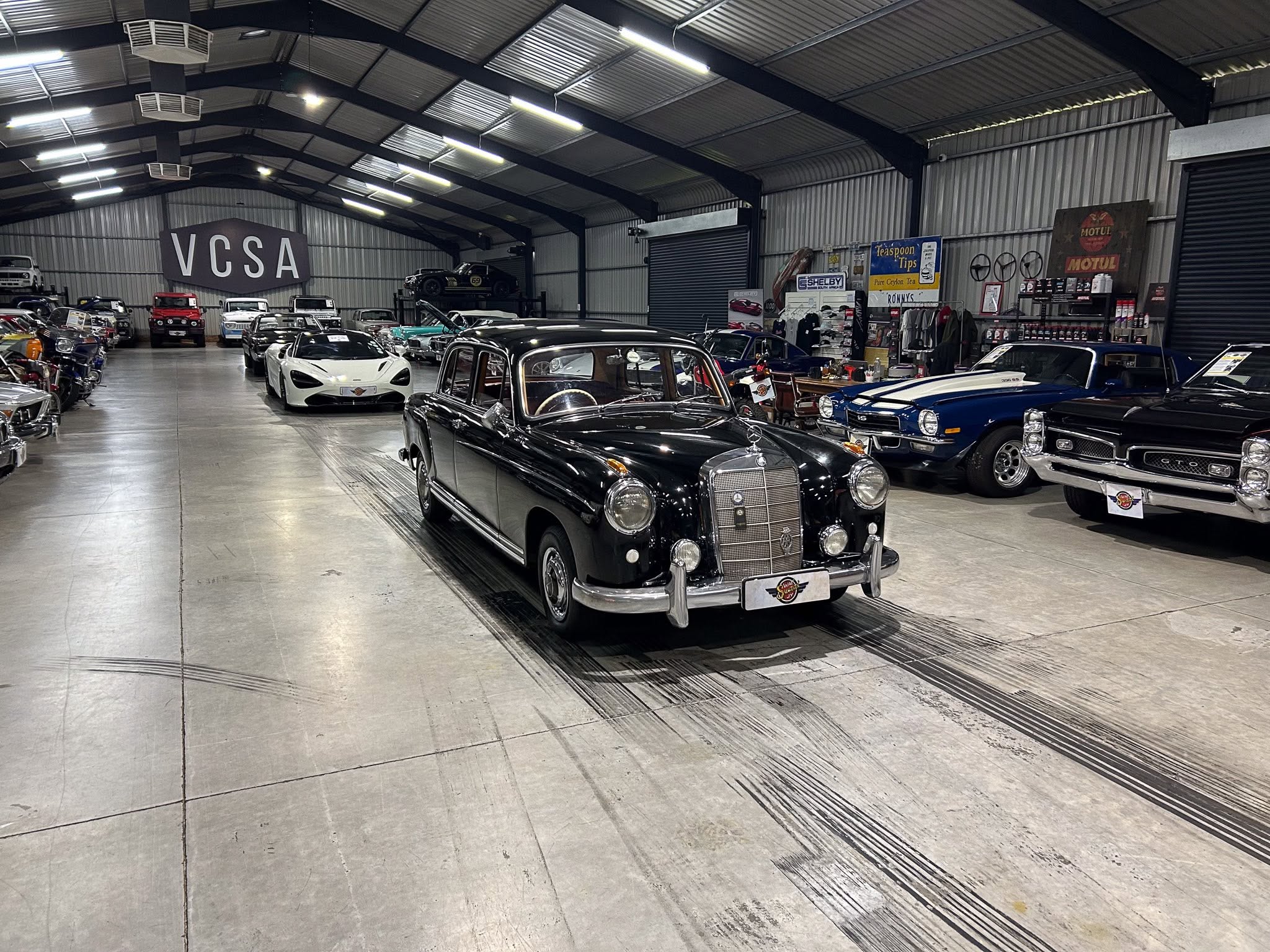 Indoor car showroom with a row of vintage and modern cars under bright overhead lights, a large VCSA sign on the back wall.