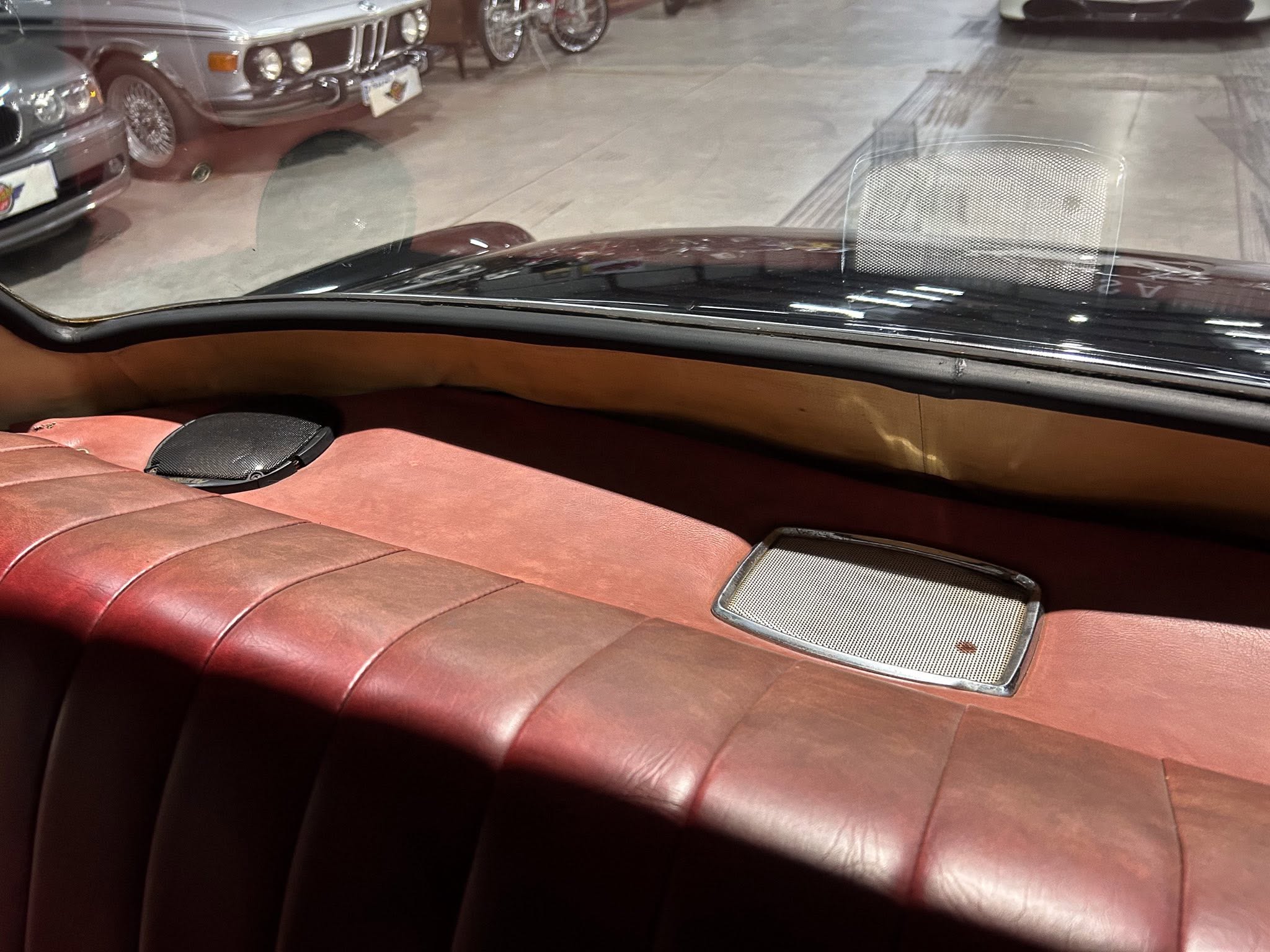 Red leather car backseat with a built-in speaker grille and chrome trim, seen through a showroom window reflecting classic cars outside
