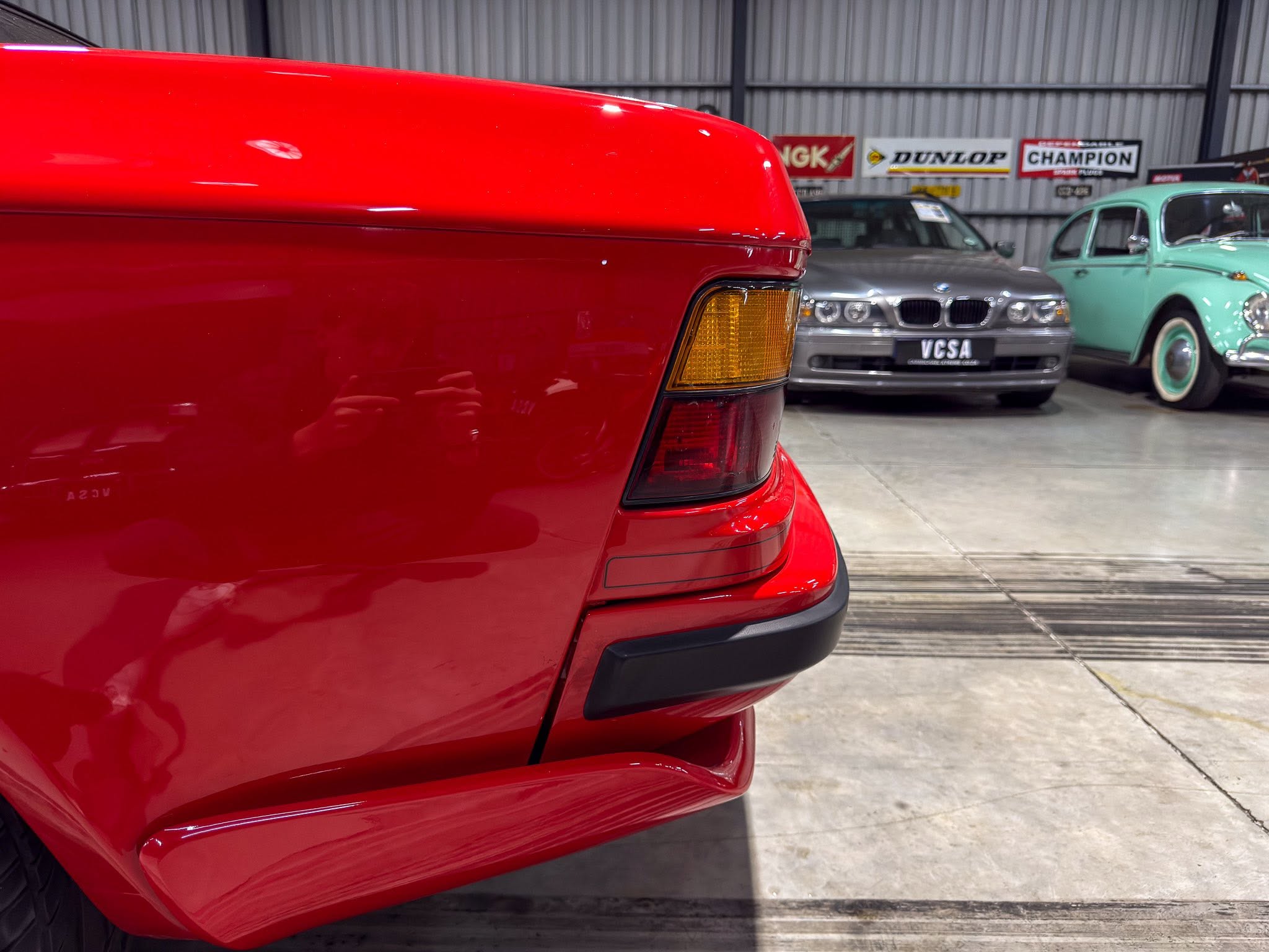 Close-up of the rear end of a glossy red car, showing the taillight and bumper in a showroom with other vintage cars in the background.