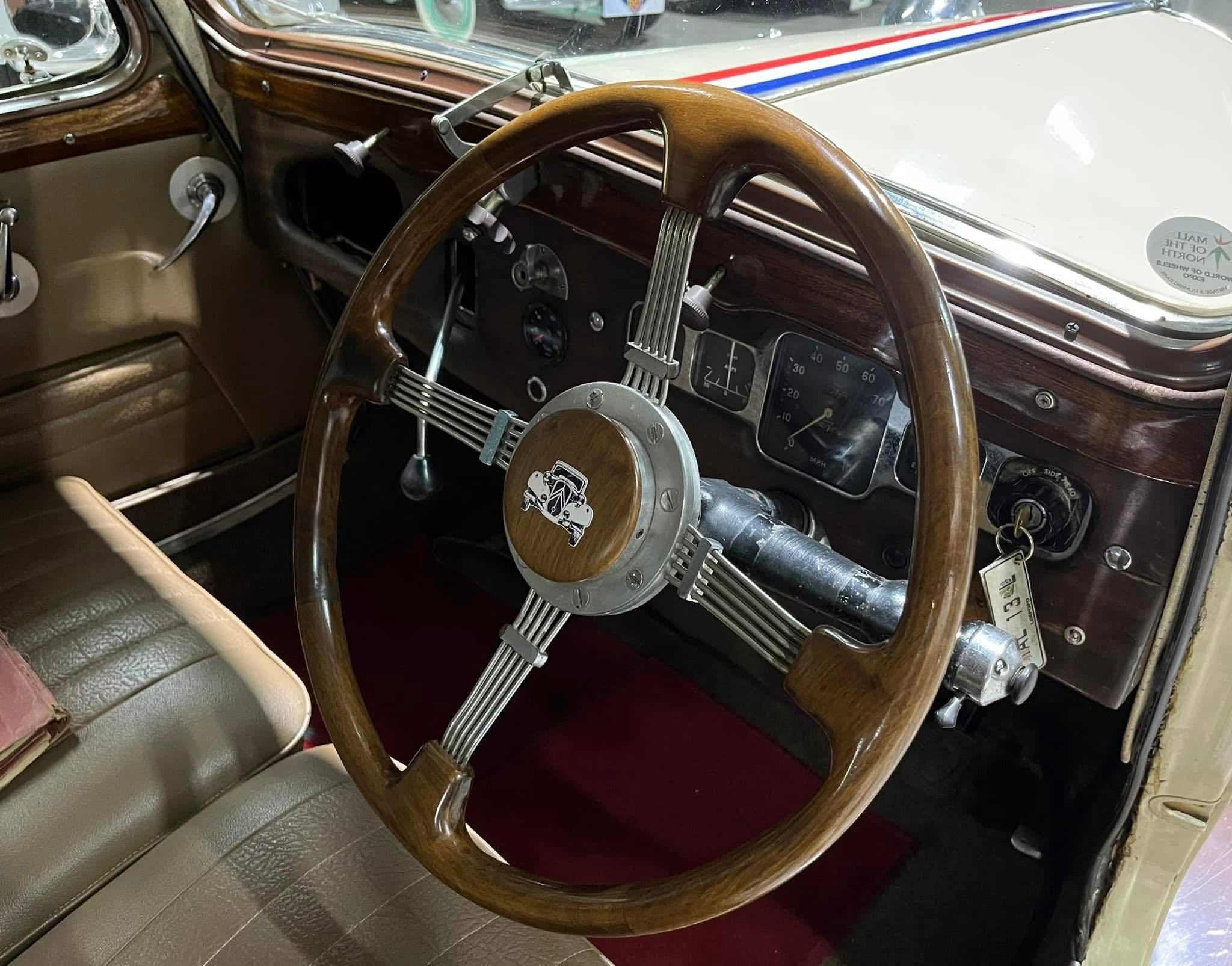 Vintage car interior with a large wooden steering wheel and chrome accents; brown leather seats and classic dashboard gauges visible.