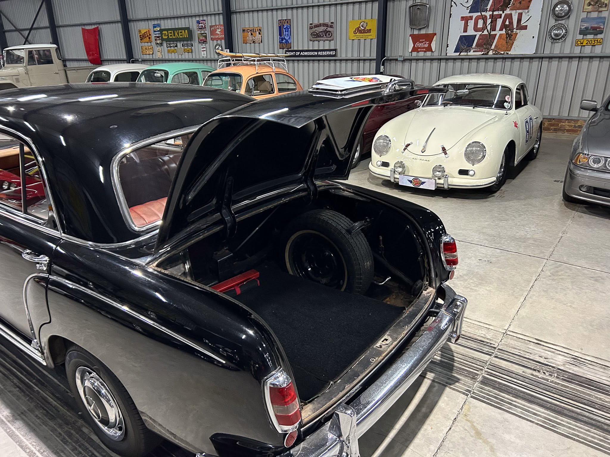 Black classic car with open trunk in a showroom of vintage vehicles; other old cars line the background.