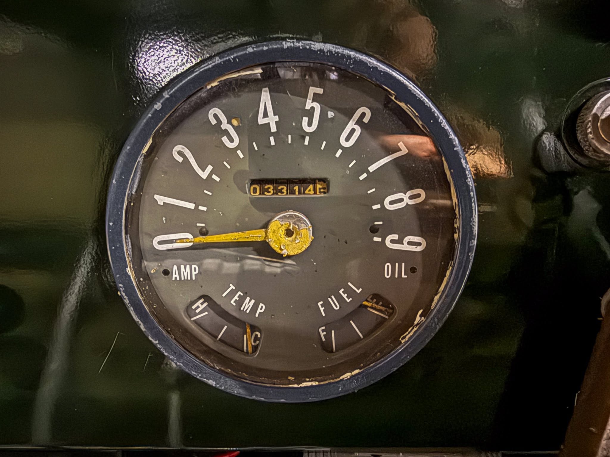Close-up of a vintage round engine gauge with a yellow needle; numbers 0–8 around the dial and an odometer reading 03314, with AMP and OIL labels visible.