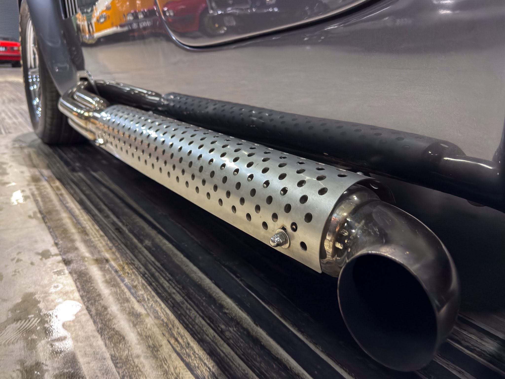 Dark vehicle side with perforated metal running board and visible exhaust pipe near the rear wheel, showing a rugged step surface.