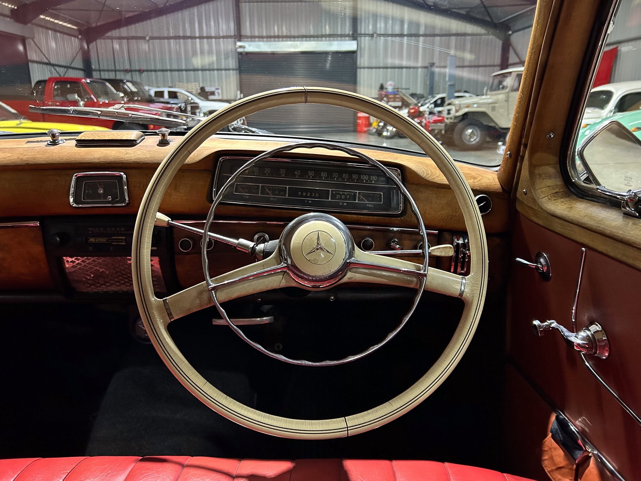 View from the driver’s seat of a vintage car: large cream steering wheel, wood dashboard, chrome accents, and red leather seating, inside a showroom with other classic cars in the background.