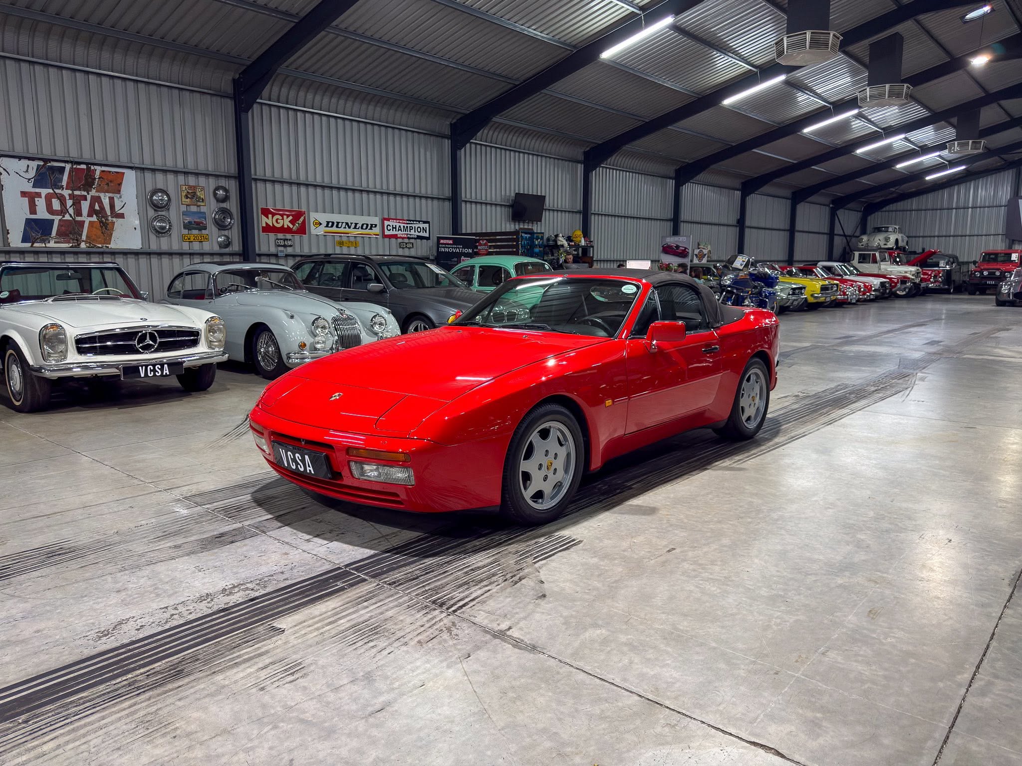 Indoor showroom of a classic car collection with a bright red convertible in the foreground and vintage cars along the wall behind it.