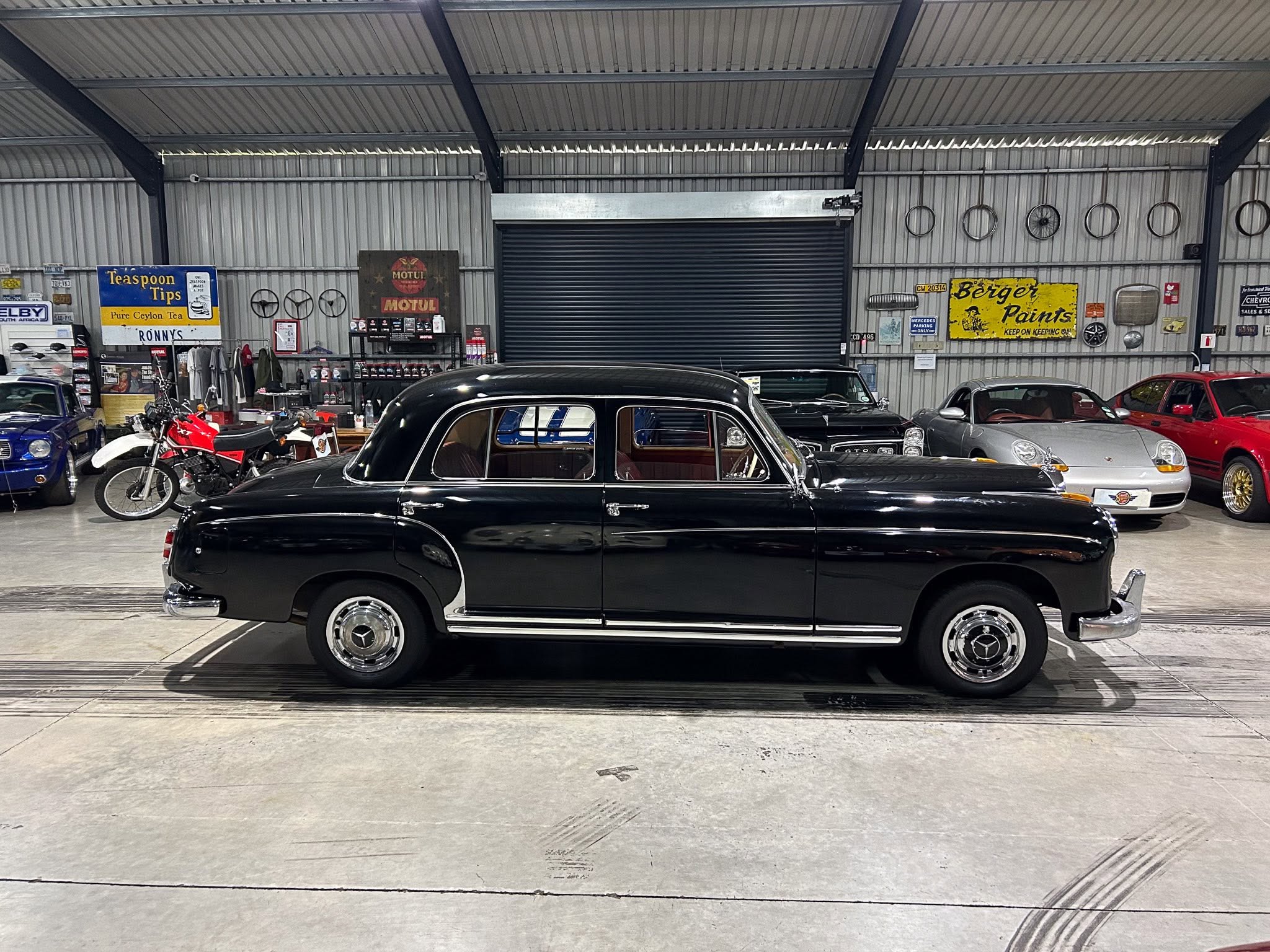 Indoor classic car showroom with a glossy black vintage sedan in the foreground and other vintage cars in the background; wall displays include bicycle wheels and signage like Motul and Bergér Paints.