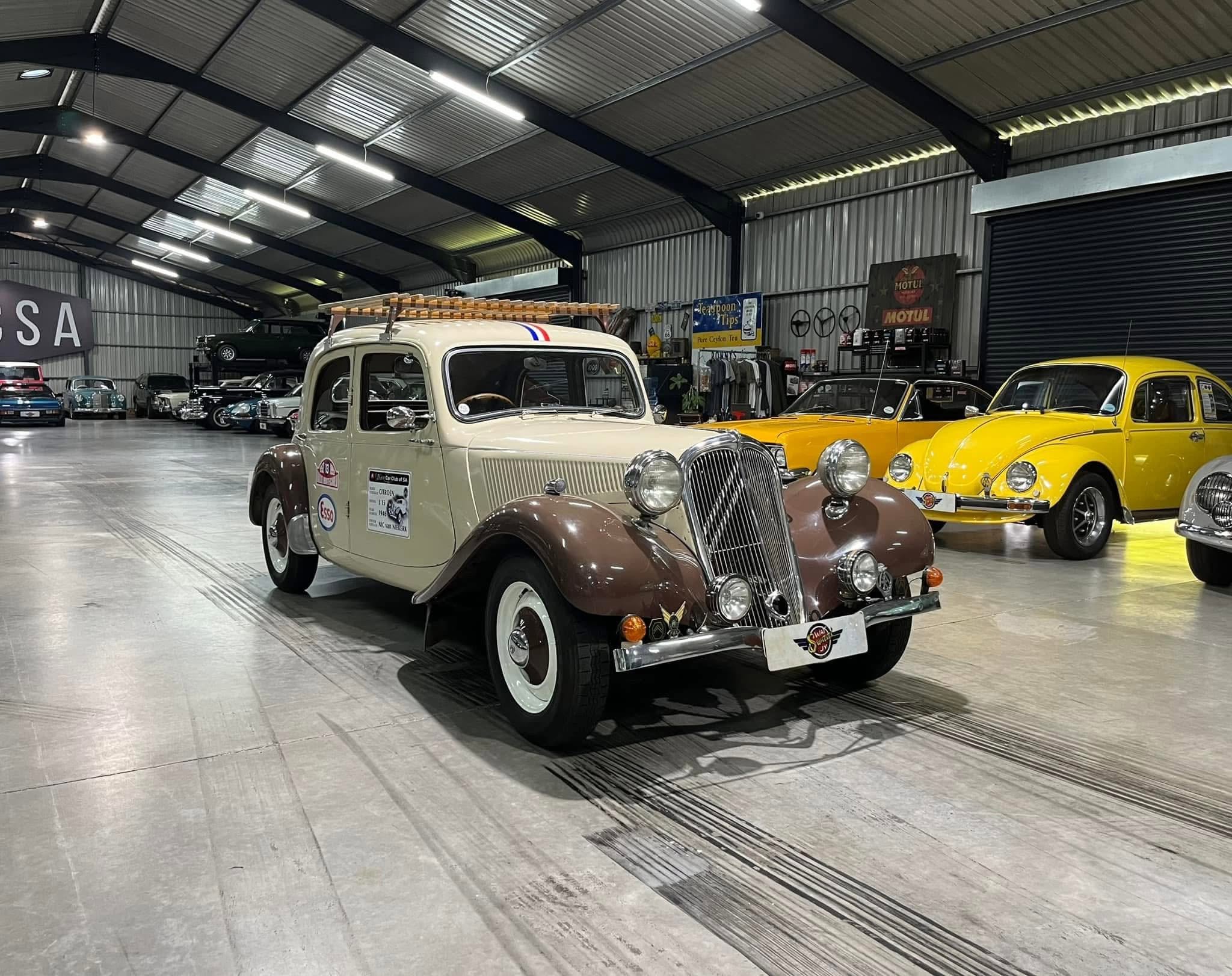 Indoor showroom displaying vintage cars; a cream-and-brown classic in front with yellow cars behind it under a metal-roofed ceiling.