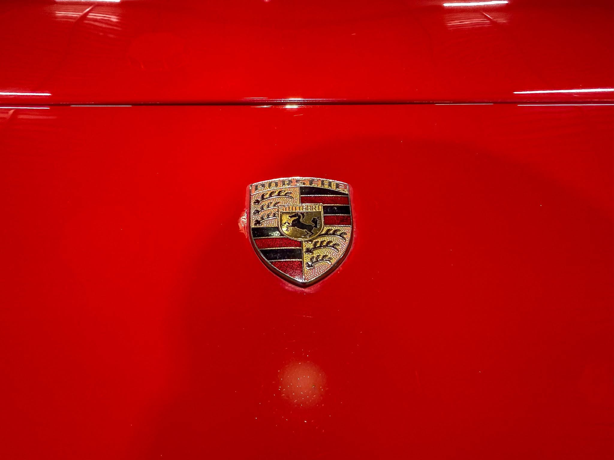 Close-up of a Porsche crest badge on a glossy red car hood with a chrome seam above the emblem