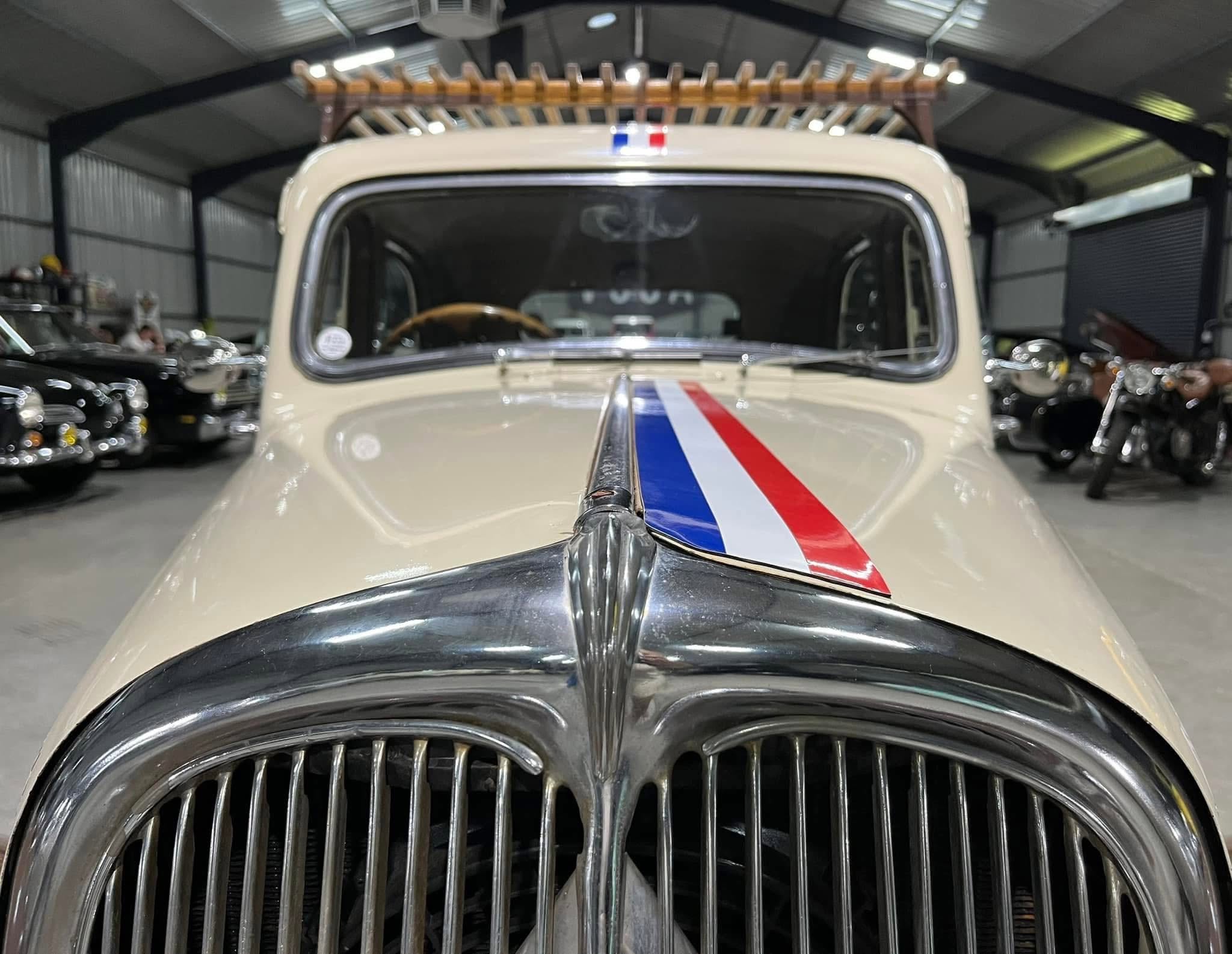 Front view of a vintage car inside a garage, beige body with red, white, and blue racing stripe along the hood, chrome grille visible.