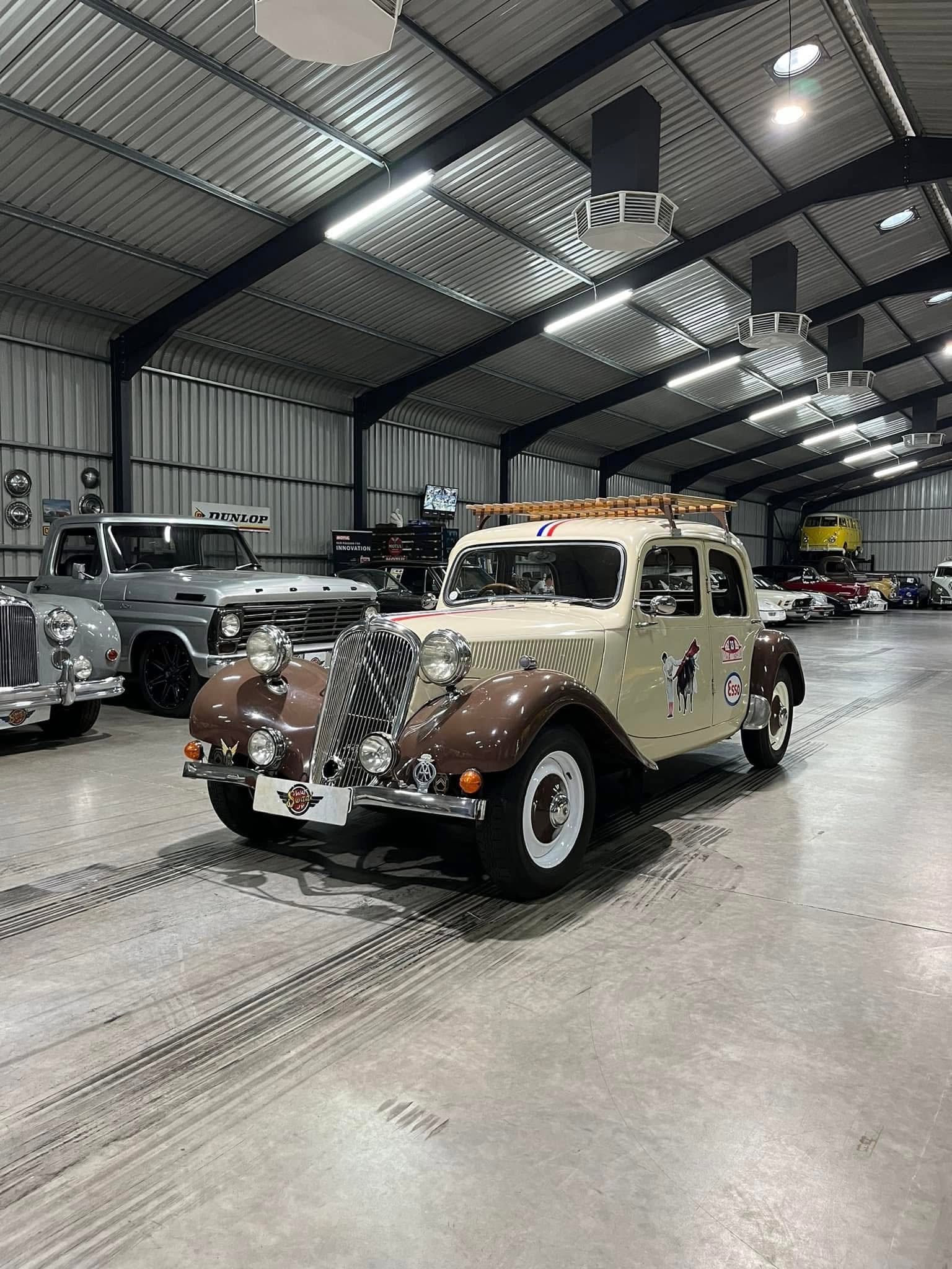 Cream and brown vintage car in a bright industrial-style showroom with other classic cars in the background and steel beams overhead.