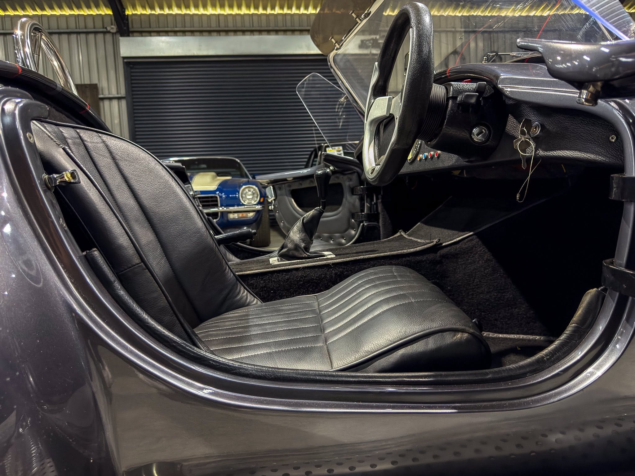 Interior of a classic black car with a right-hand steering wheel, black leather seats, and keys in the dashboard ignition; a blue car is visible in the background inside a garage.