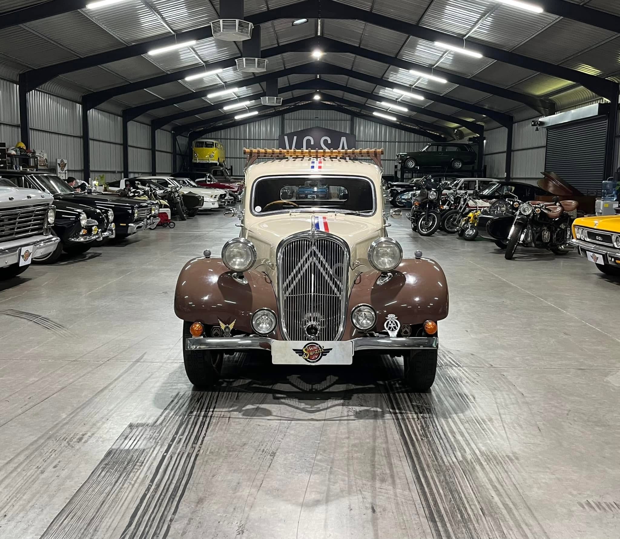Indoor showroom for classic cars, with a beige vintage car centered in the foreground and rows of other classic vehicles lining both sides of the space.