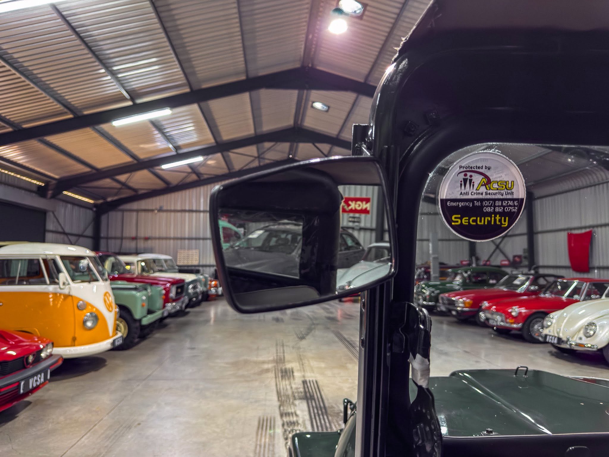 Interior view of a warehouse-style showroom filled with colorful classic cars, seen through a car's side mirror
