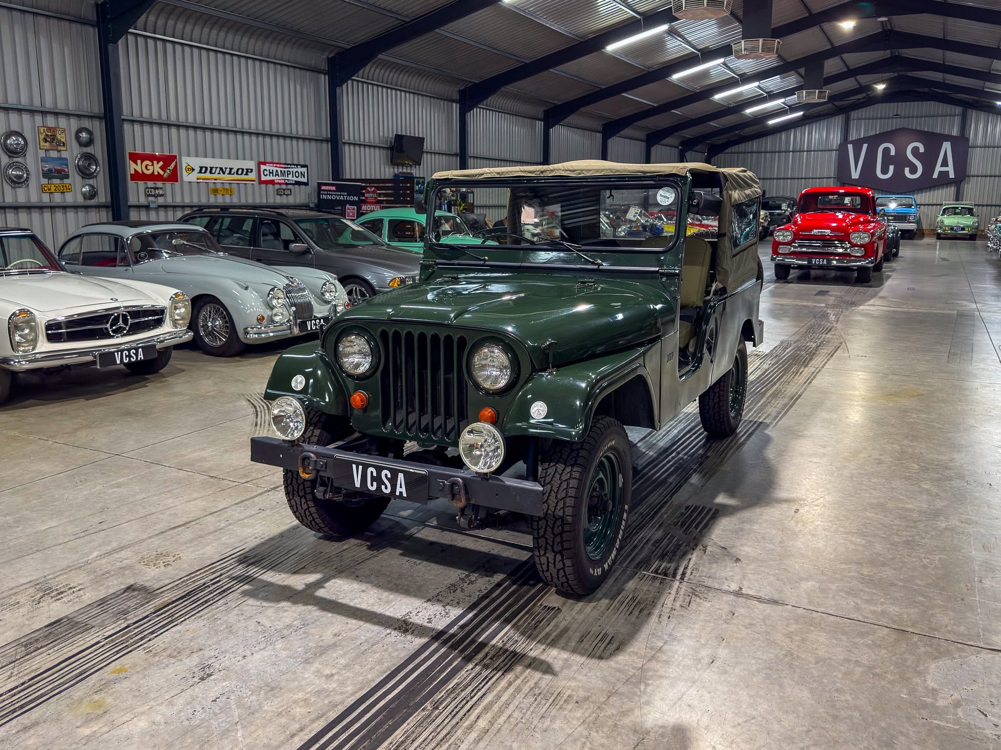 Indoor classic car showroom with a green jeep in the foreground and vintage cars lined along steel walls.