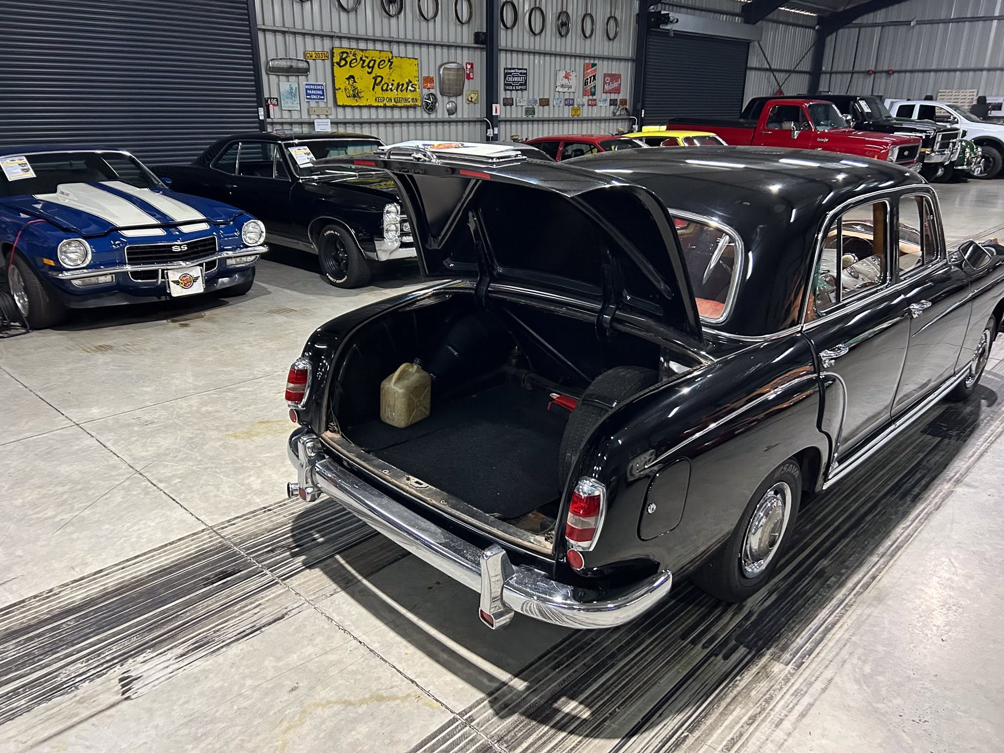 Indoor showroom of classic cars with a black vintage sedan trunk open in the foreground and other polished cars parked along the walls, signs on the corrugated metal wall behind.