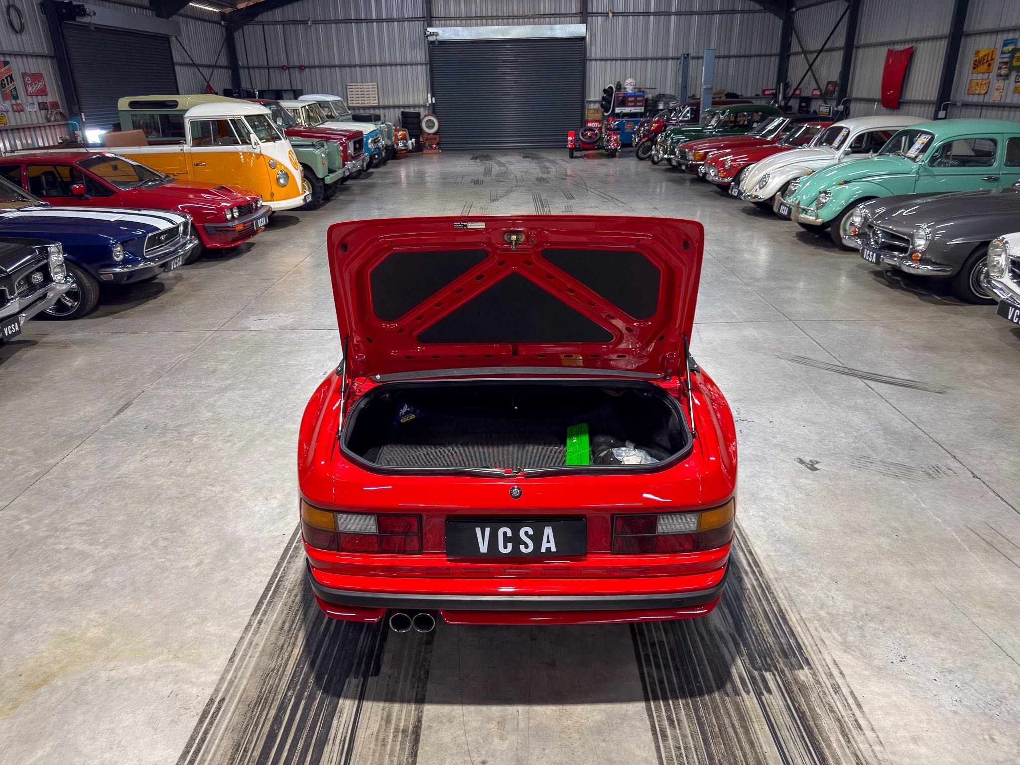 Red hatchback with the trunk open, seen from the rear, in a warehouse showroom lined with vintage cars on both sides.