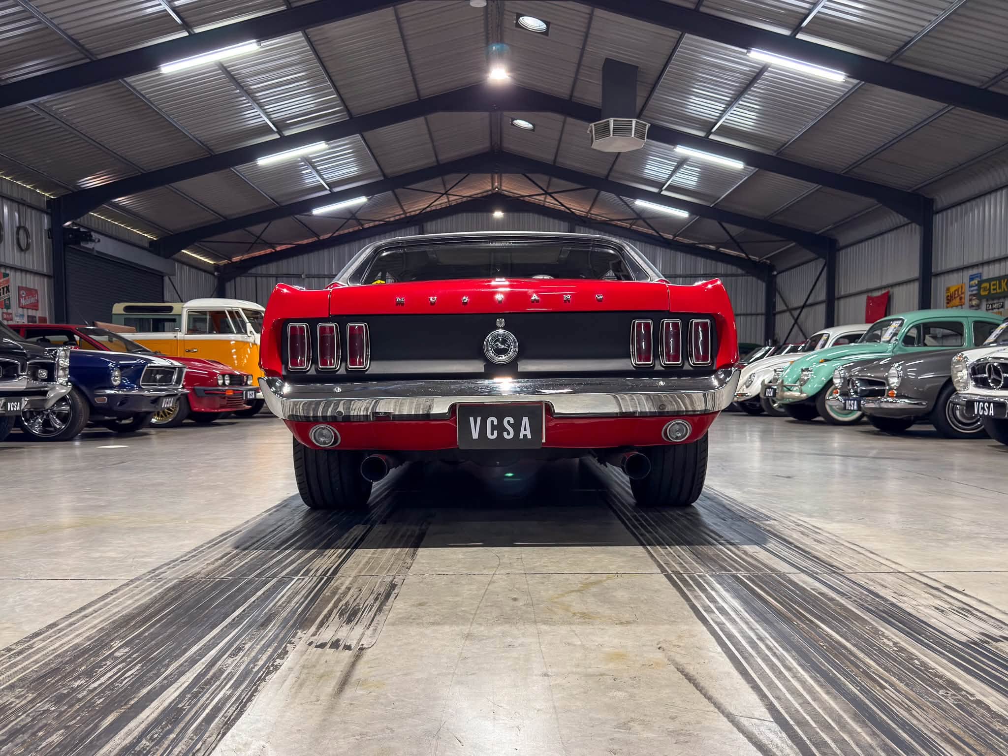 Red Ford Mustang seen from behind in a spacious classic car showroom filled with vintage vehicles