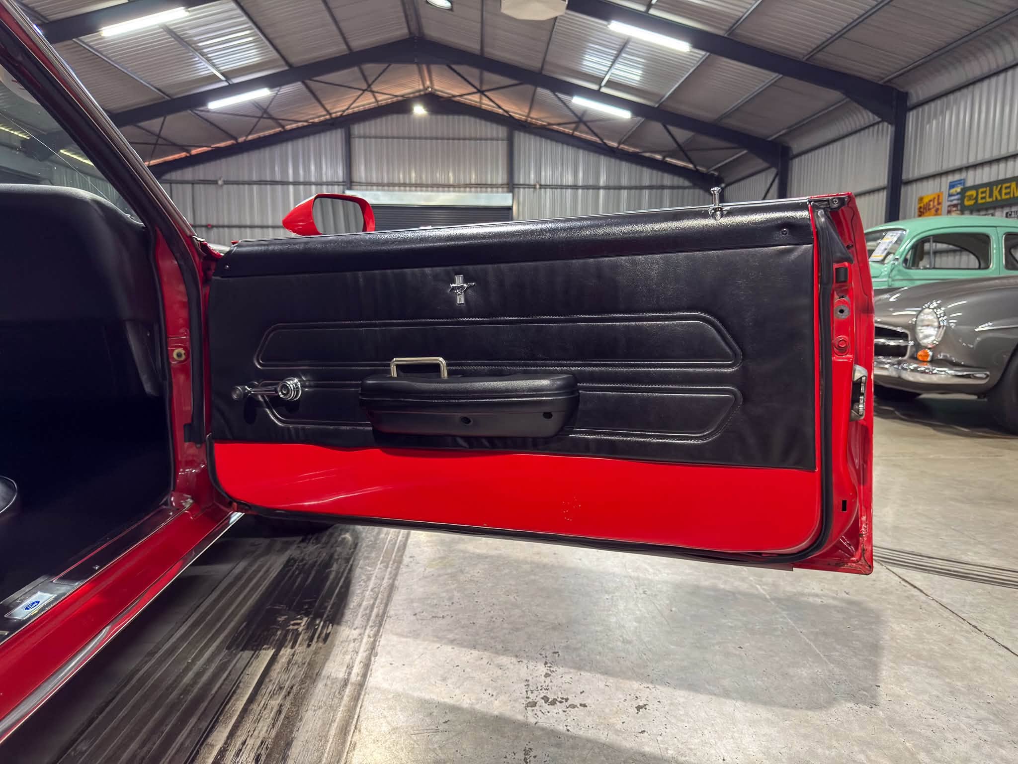 Red car door open to reveal black interior with a Mustang emblem, inside a metal-structured garage with vintage cars in the background