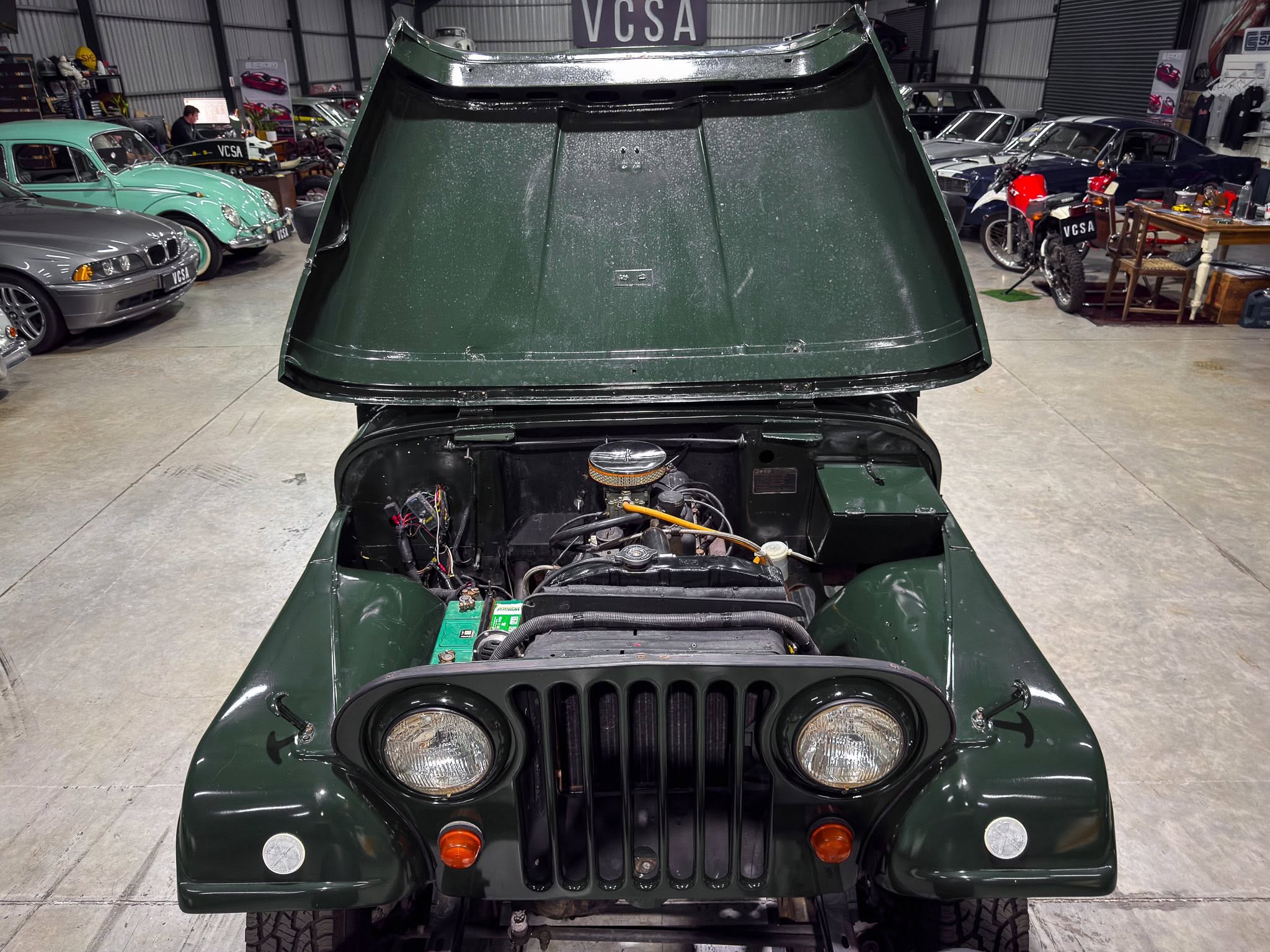 Dark green Jeep with its hood up in a garage, engine visible and other classic cars in the background in a showroom.